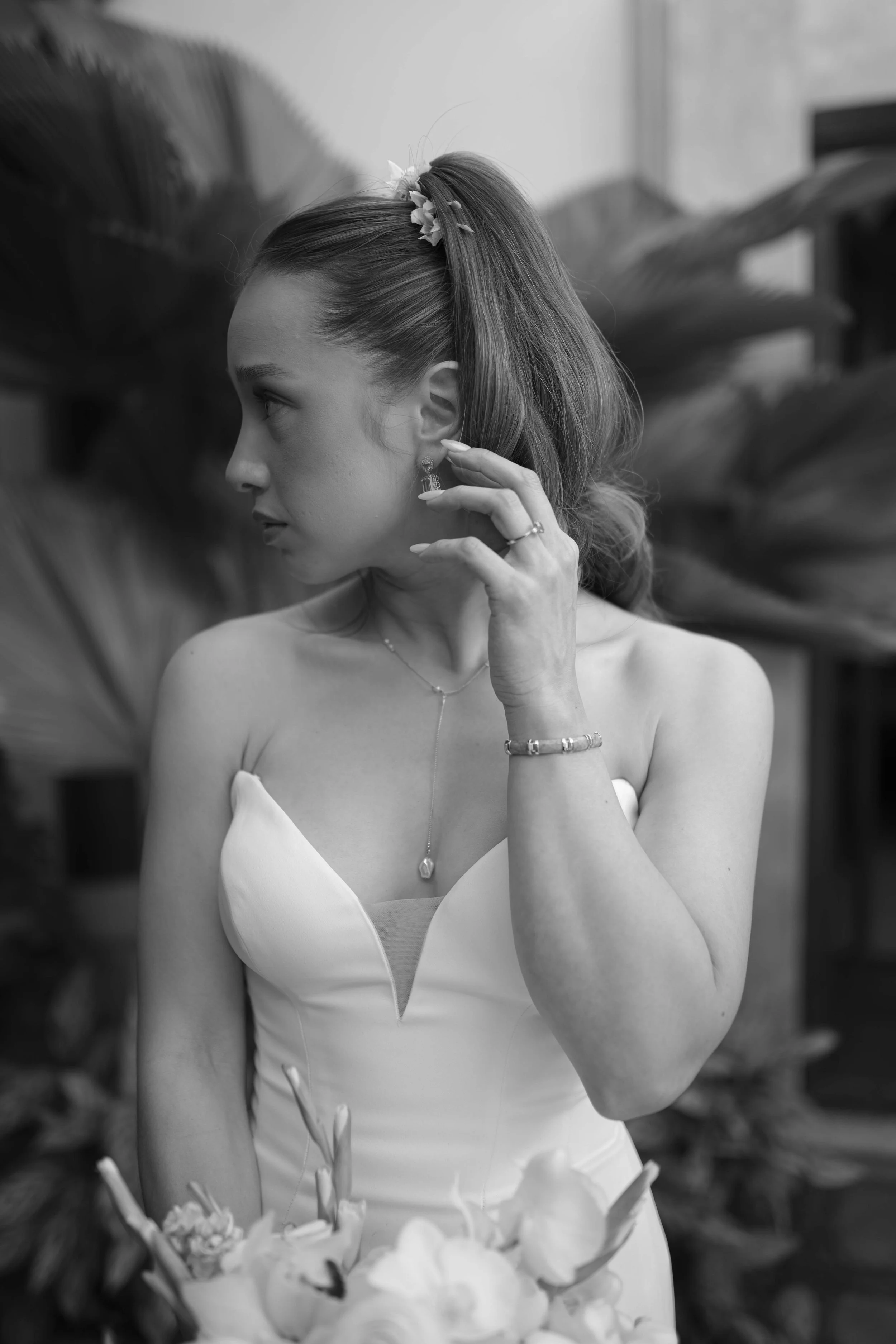 Retrato en blanco y negro de una mujer con vestido de novia sosteniendo un ramo de flores, mirando hacia la izquierda, con accesorios de joyería y un peinado con flores, en un fondo de plantas tropicales.