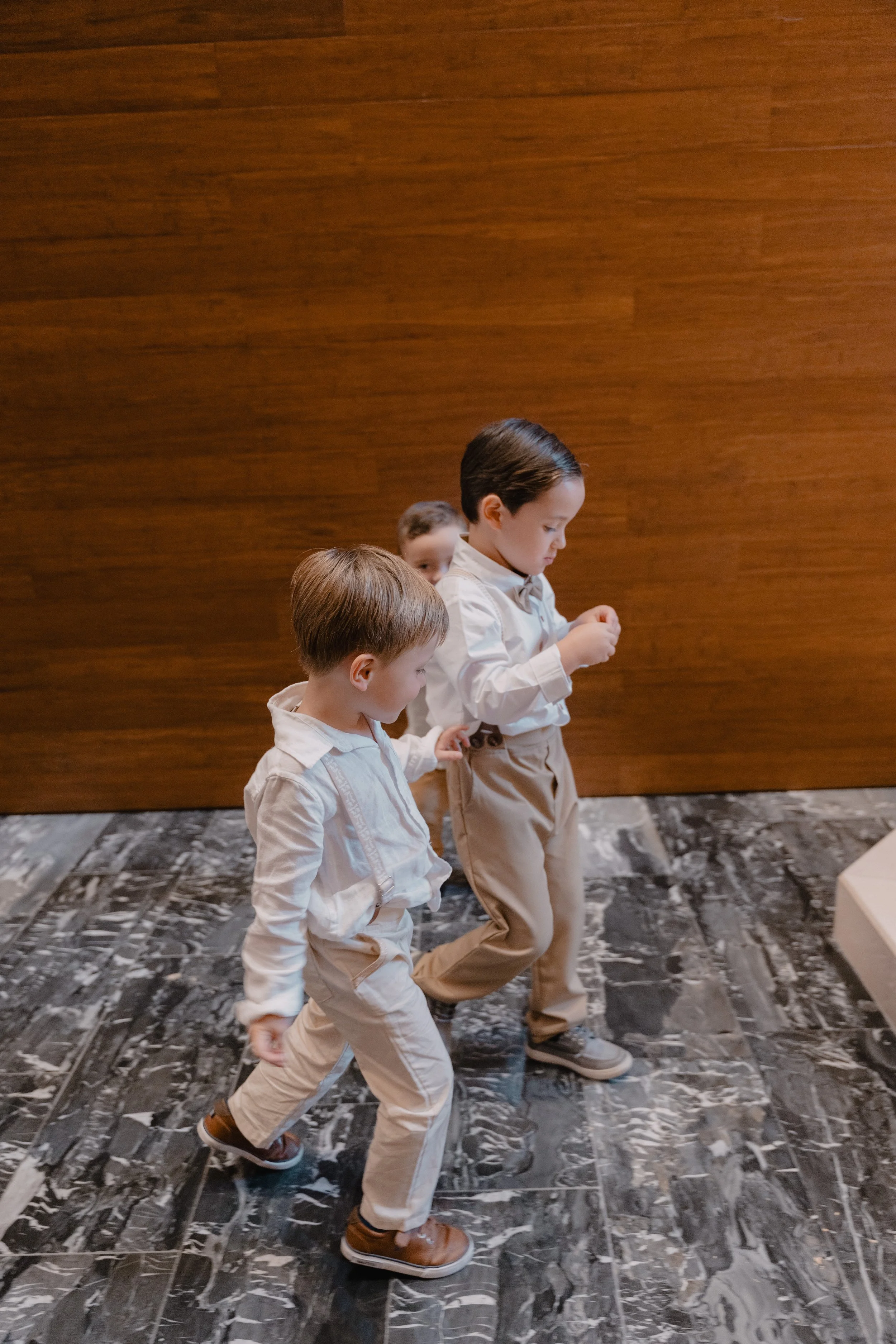 Tres niños pequeños caminando en un interior con suelo de mármol y pared de madera.