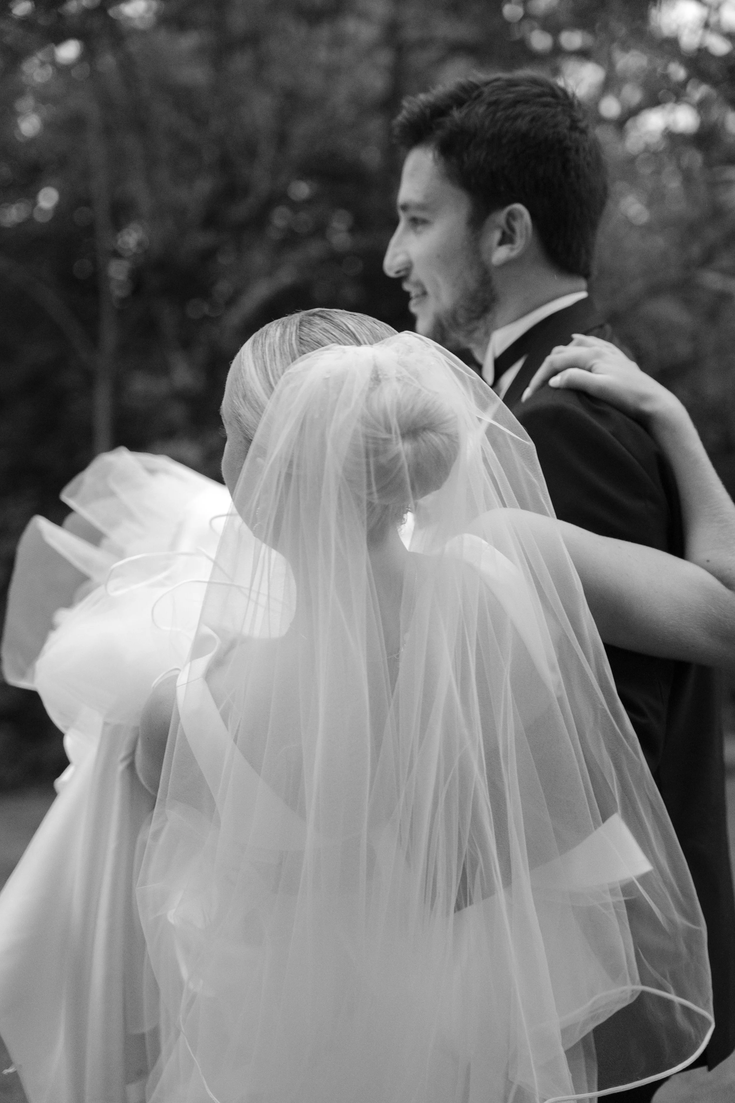 Pareja en boda, la mujer con velo y vestido, el hombre con traje, abrazándose en un fondo de árboles.