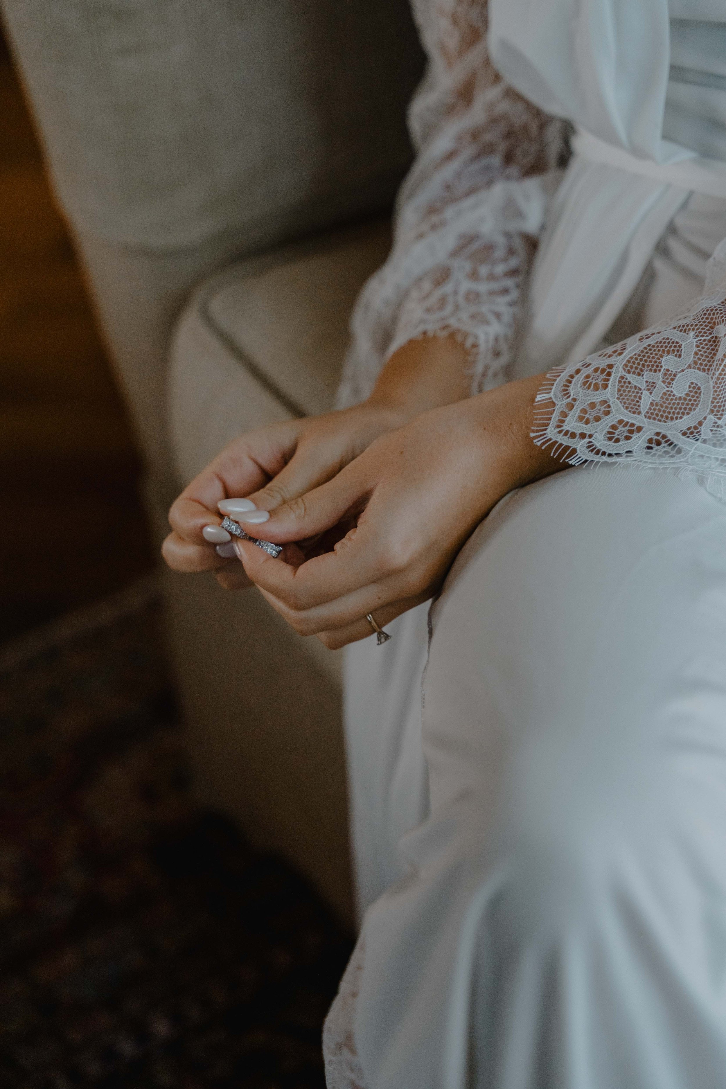 Manos de una mujer en vestido de novia sosteniendo un anillo en su mano, sentada en una silla.