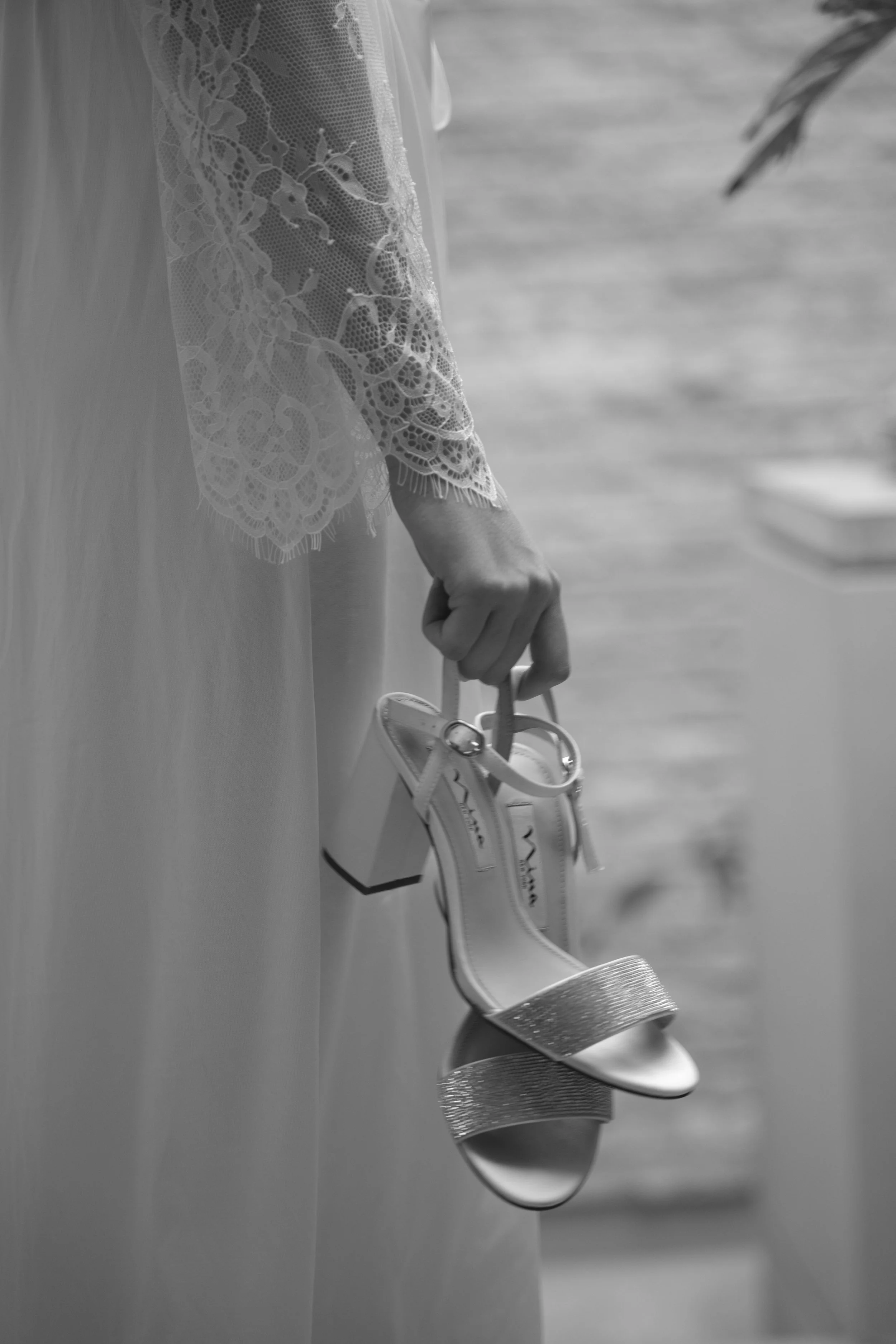 Una mano sostiene unos zapatos de tacón altos de estilo elegante, con detalles brillantes, mientras que una persona viste un vestido de encaje y seda en un entorno que parece ser cerca del agua.