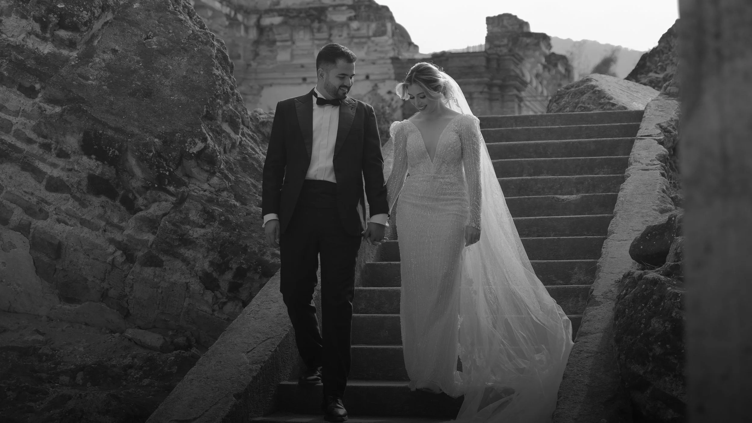 Pareja de recién casados bajando unas escaleras de piedra en un lugar histórico o al aire libre, ella con vestido de novia largo y él en traje formal. La imagen es en blanco y negro.