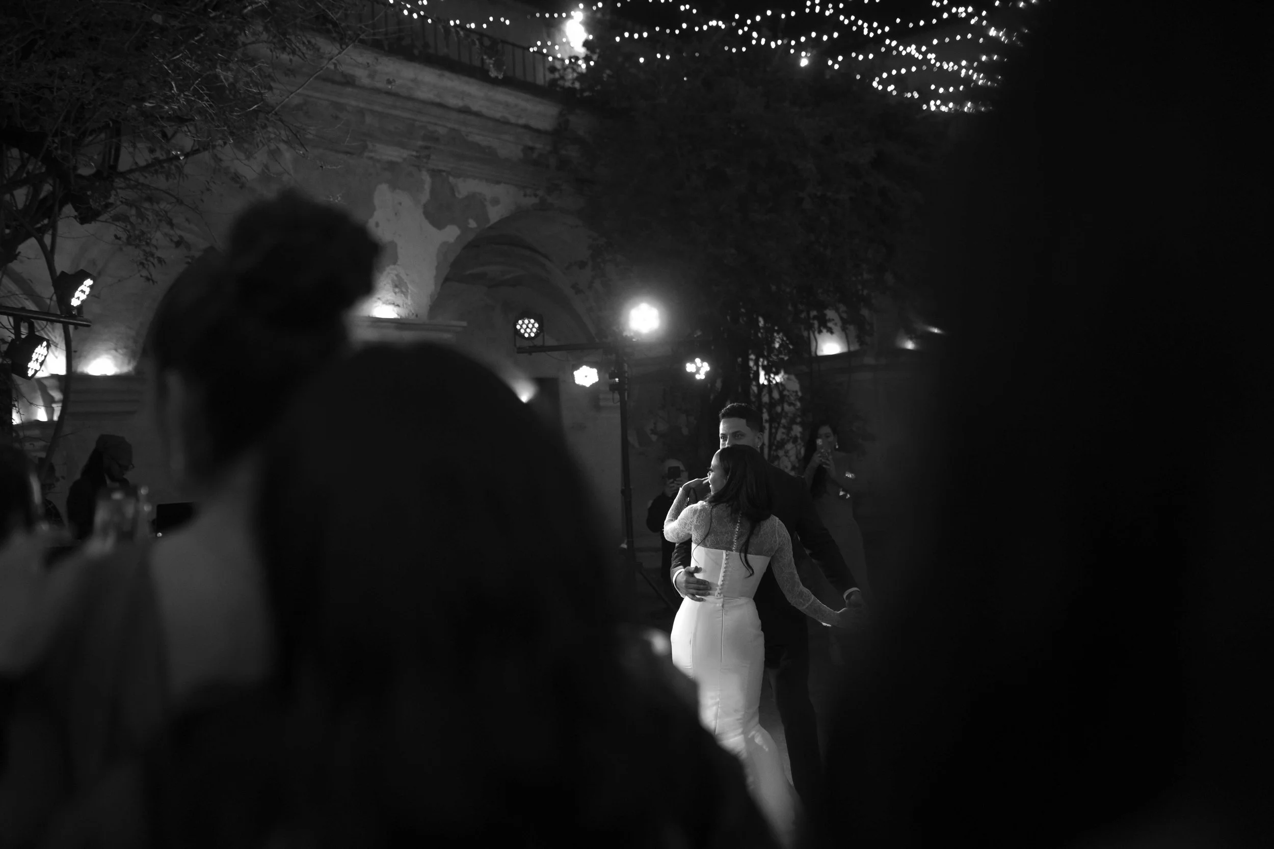 Pareja bailando en una ceremonia de boda en un lugar decorado con luces y arcos, en un entorno oscuro con personas observando.