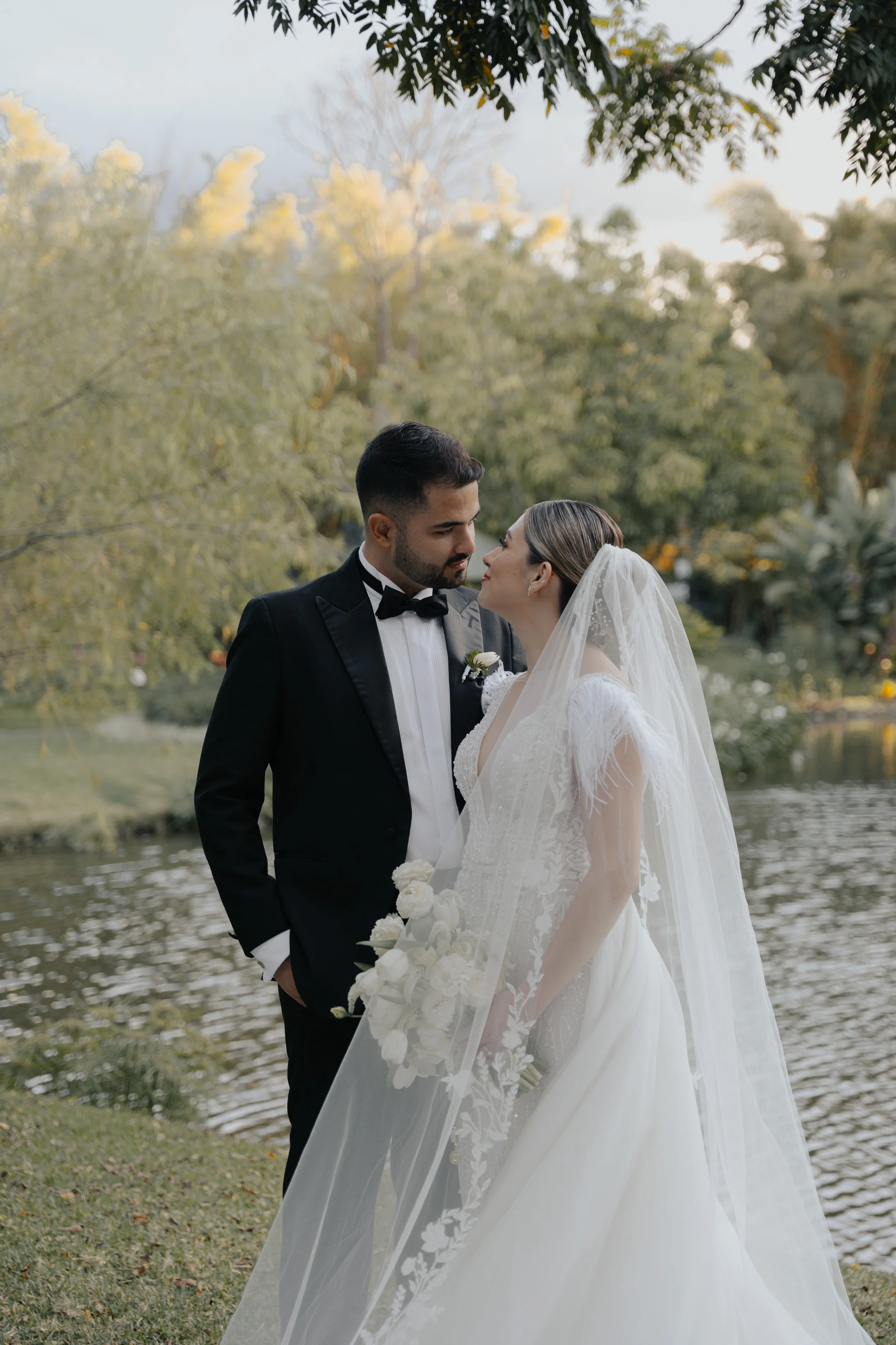 Pareja de novios de boda en un parque cerca de un lago, con árboles verdes en el fondo, mirándose cariñosamente.