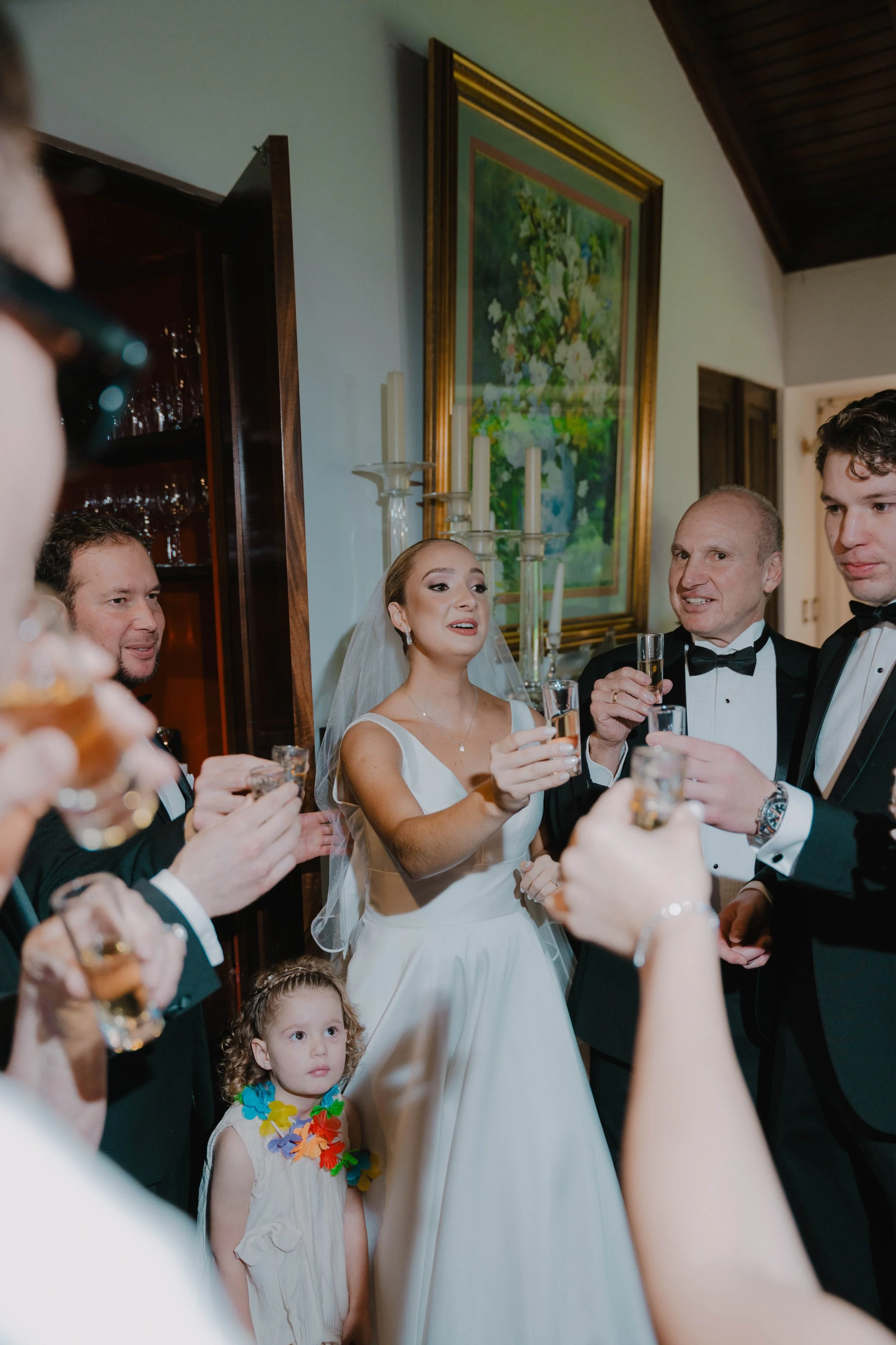 Boda: grupo de personas, incluyendo la novia y varios hombres, brindando con copas de champán en una sala decorada con cuadros y candelabros, mientras una niña observa.