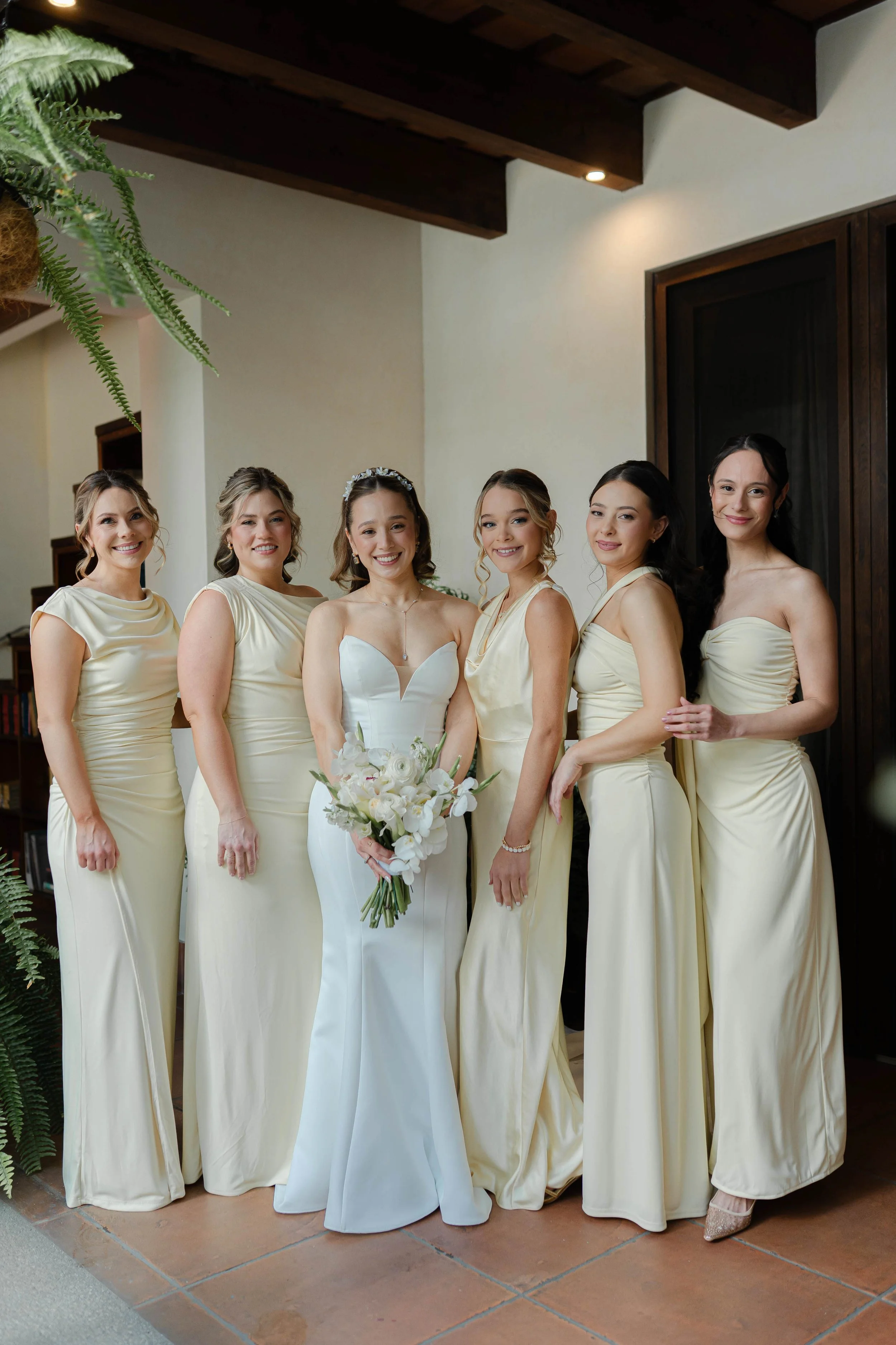 Grupo de mujeres en vestimenta de boda, incluyendo a la novia con ramo de flores, en un entorno interior con decoración moderna.