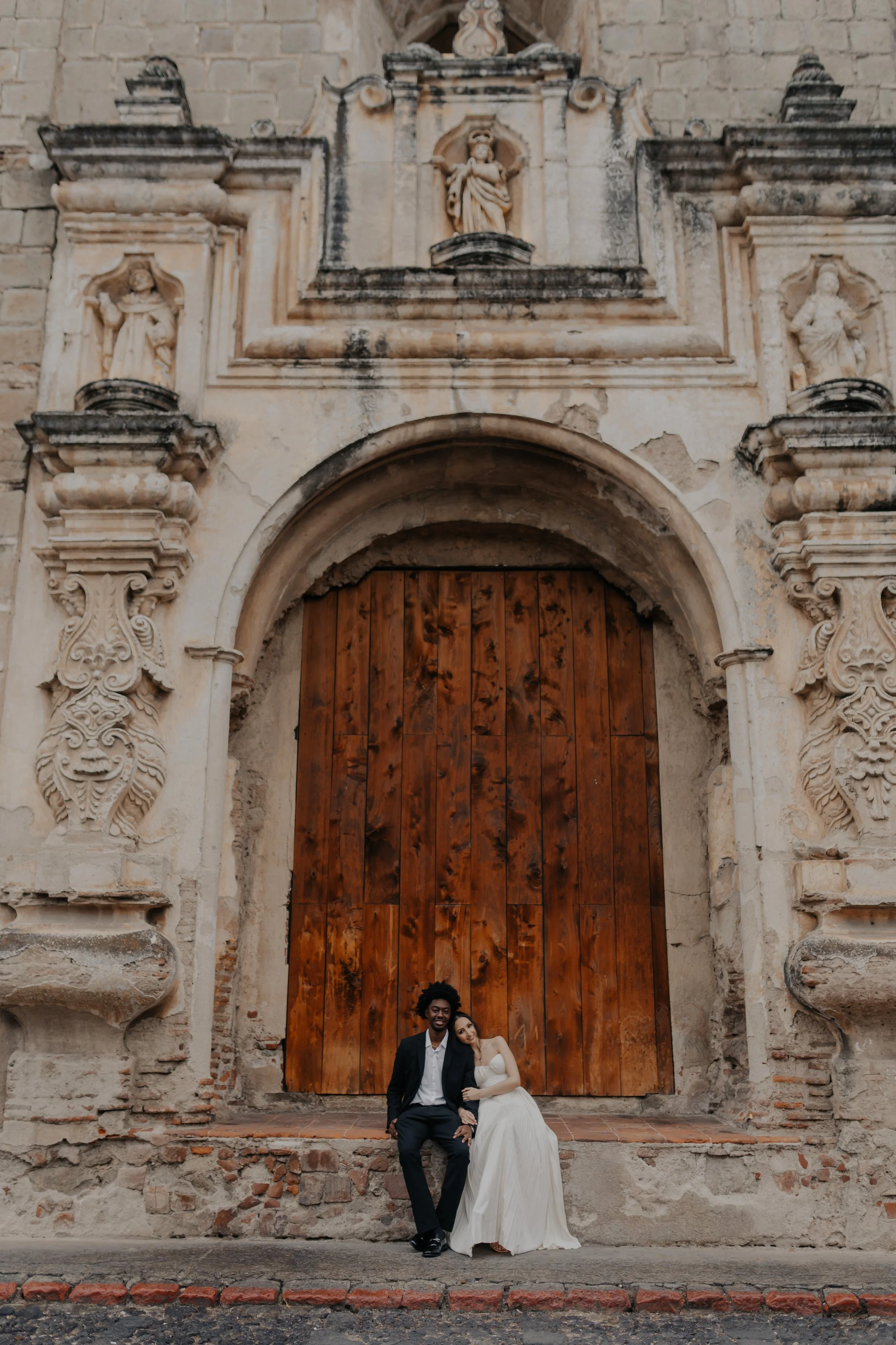 Una pareja de recién casados sentados frente a una gran puerta de madera en una iglesia antigua con detalles ornamentales en la fachada.