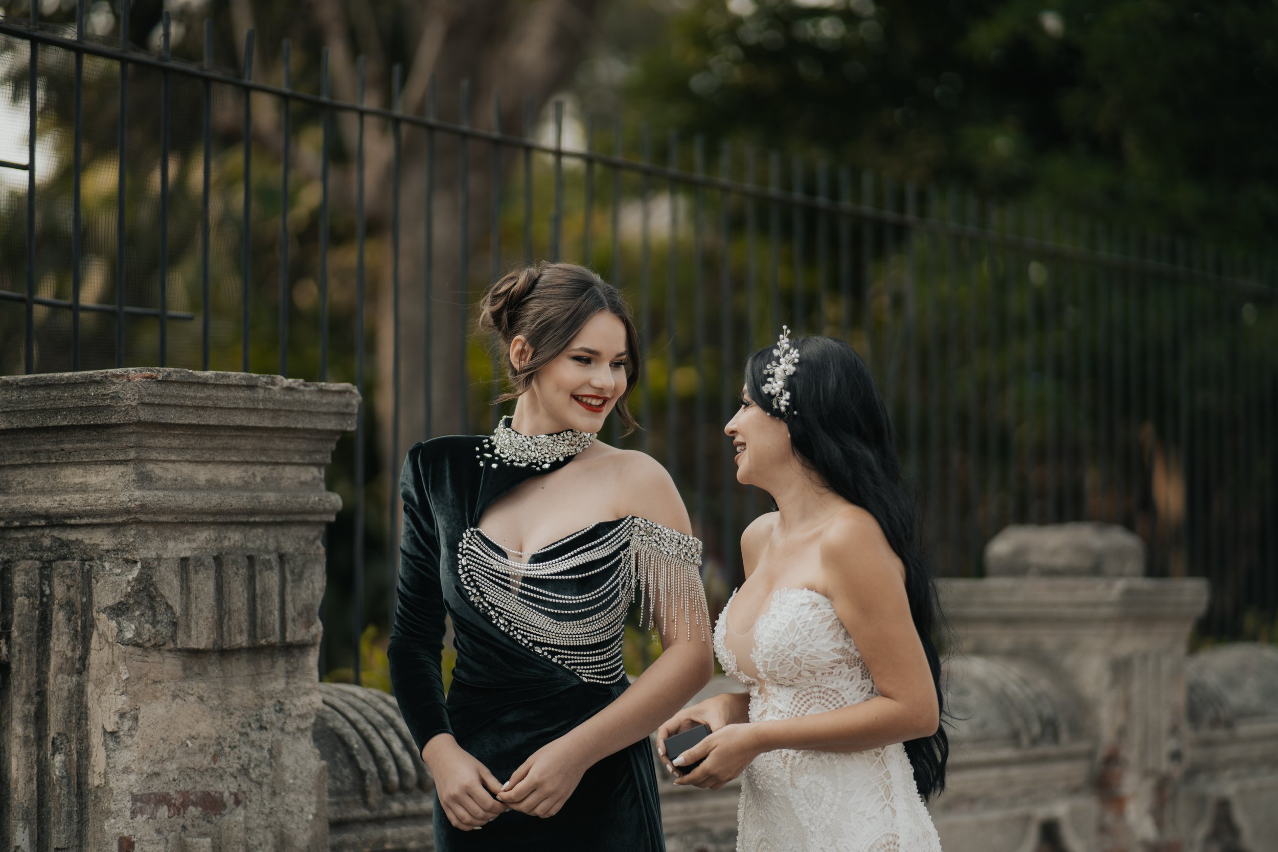 Dos mujeres vestidas con elegantes vestidos de novia y de fiesta, compartiendo una conversación en un entorno al aire libre con una cerca de hierro y arquitectura de piedra.