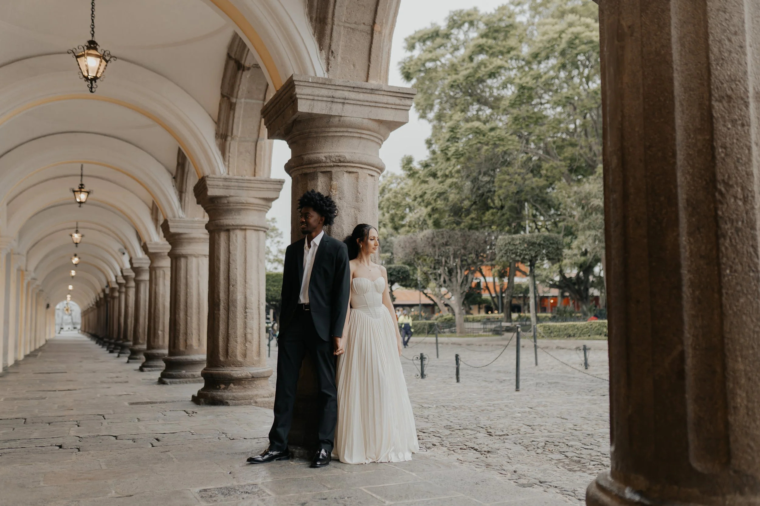Pareja de novios, ella con vestido largo blanco y él con traje negro, de pie juntos en un pasillo de arcos de piedra en un lugar histórico, con árboles y una plaza en el fondo.