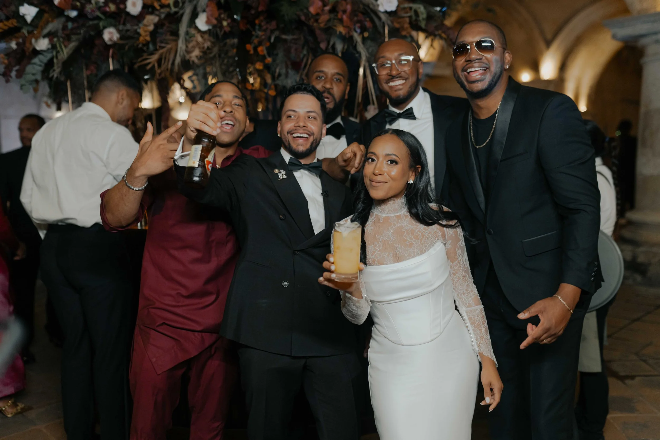 Grupo de personas en una fiesta formal, algunos con trajes de etiqueta, sonriendo y levantando bebidas en un ambiente decorado para un evento especial.