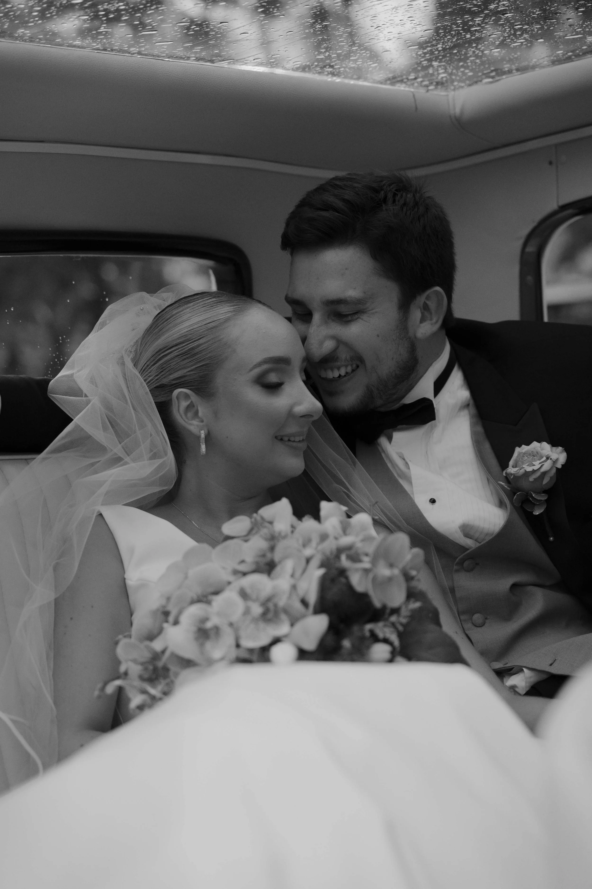 Una pareja de novios en su boda, sonriendo y acariciándose en el interior de un vehículo, la novia lleva un ramo de flores y un velo, mientras que el novio viste un esmoquin con pajarita.