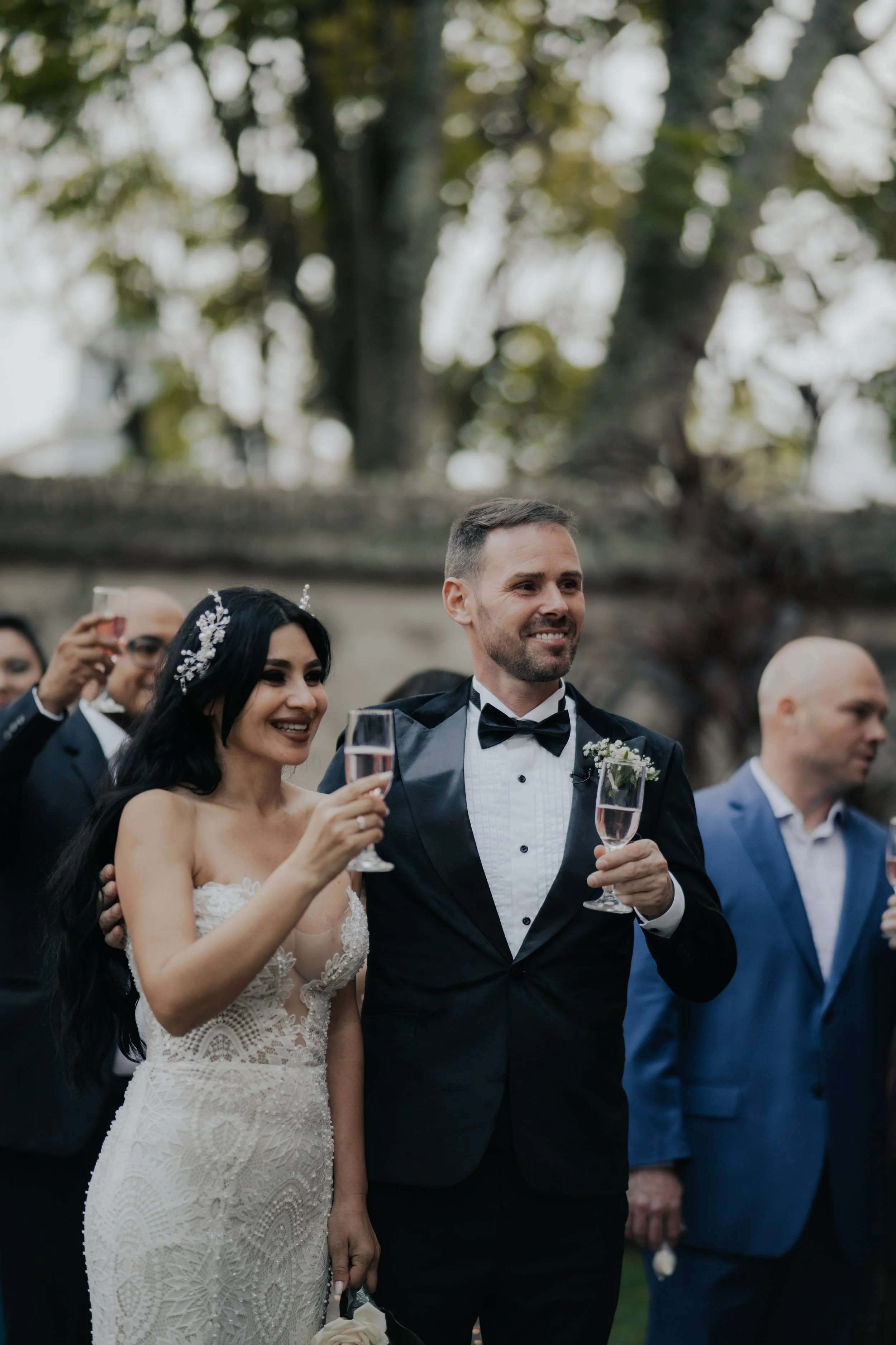 Pareja de recién casados brindando con copas de champagne en una boda al aire libre, rodeados de invitados.