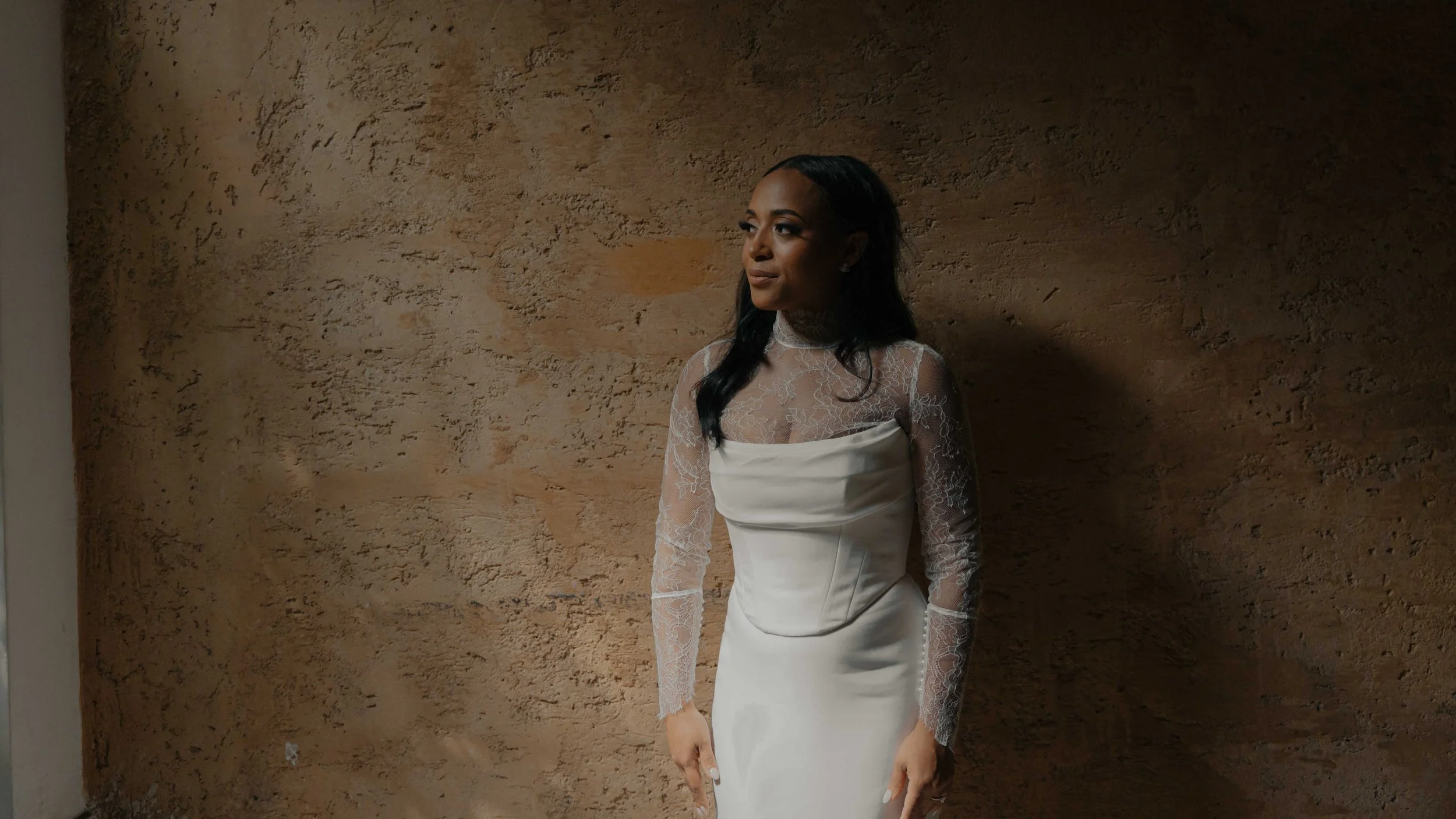 Mujer con vestido blanco y mangas de encaje, de cabello largo y oscuro, posando junto a una pared de color marrón