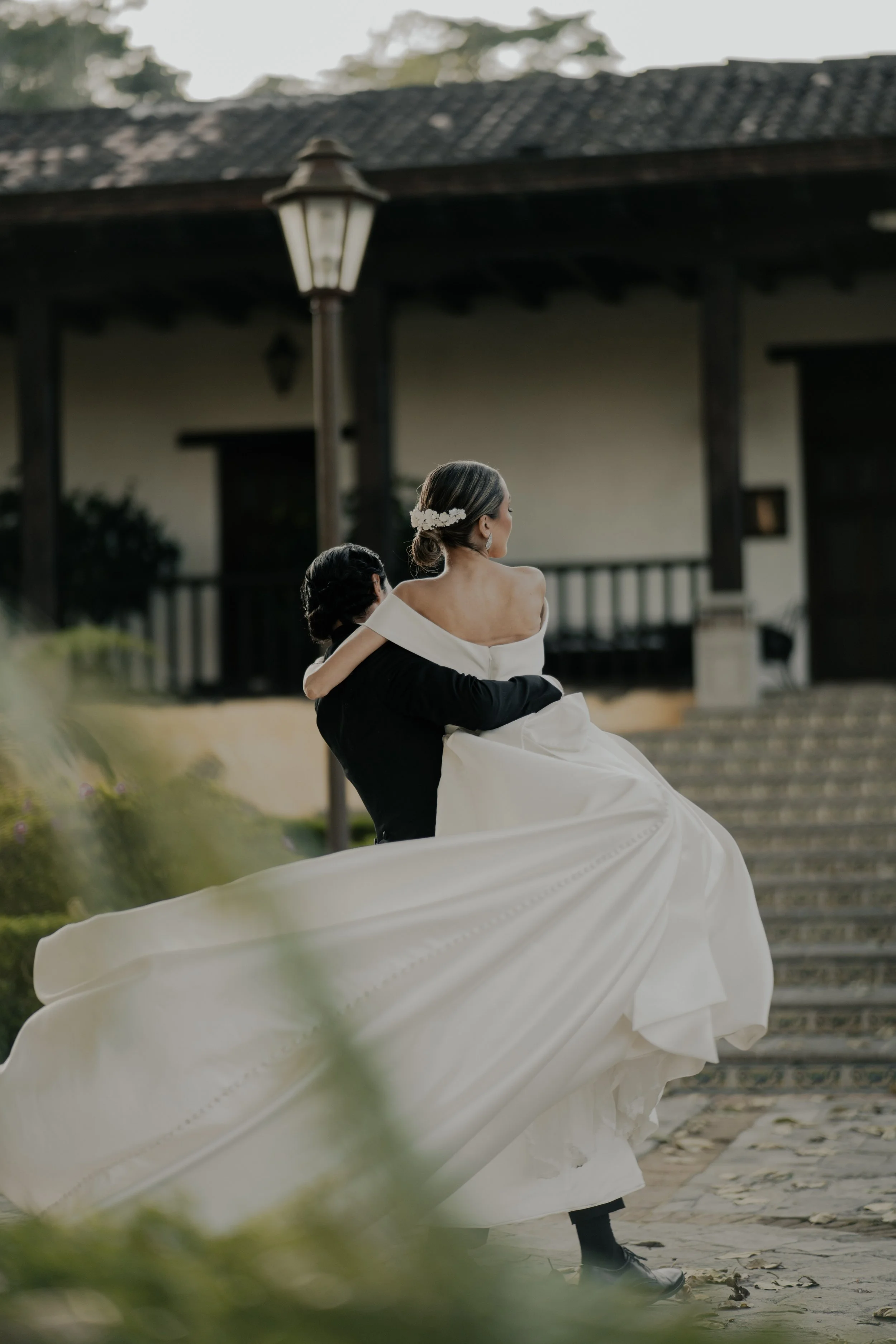 Pareja de recién casados en un paseo romántico, el novio carga a la novia en brazos frente a una casa antigua con escalones de piedra y un farol de calle.