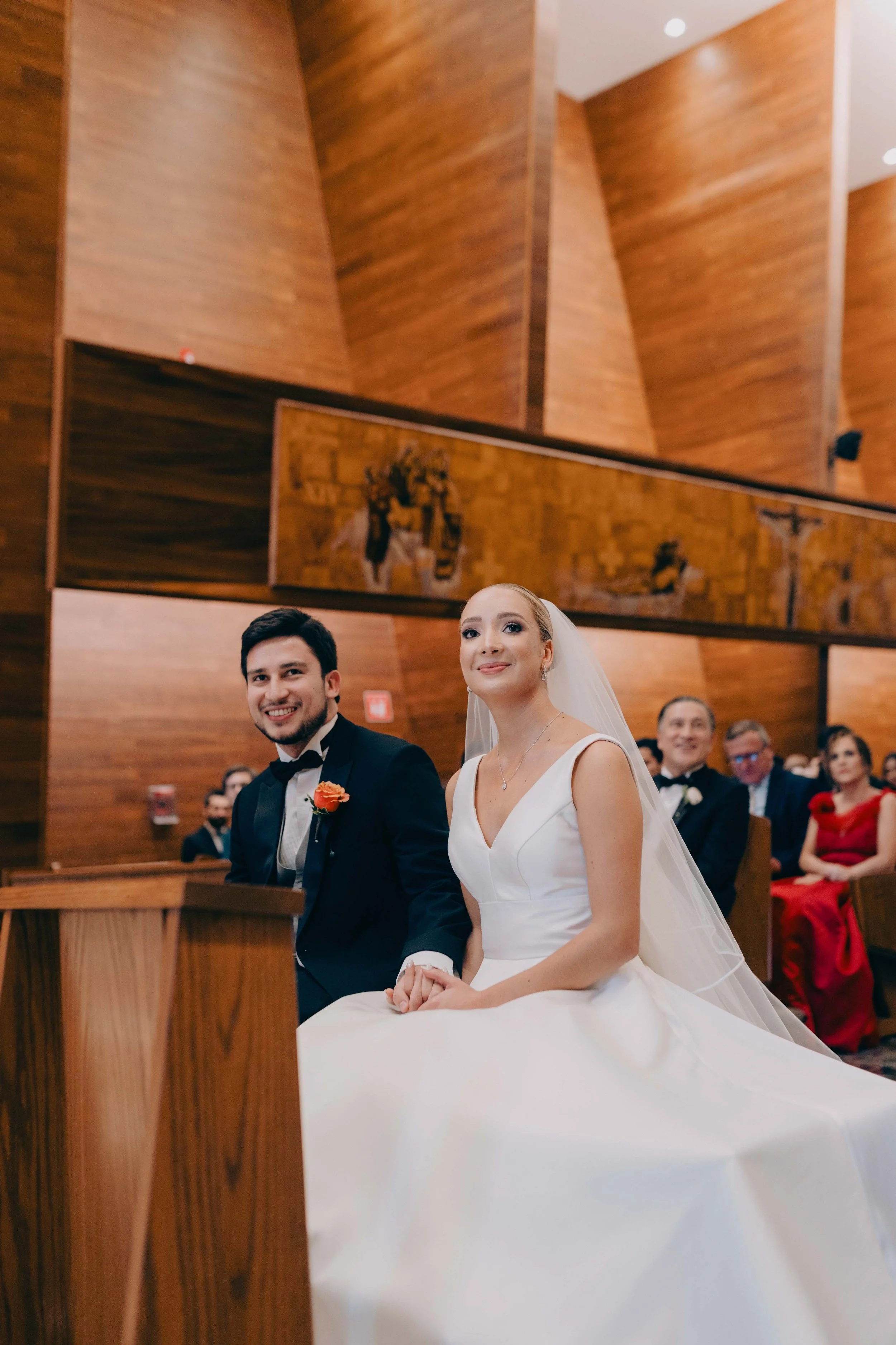 Una pareja en su boda, la novia con vestido blanco y velo, y el novio con traje negro y corbata, sentados en una iglesia de madera.
