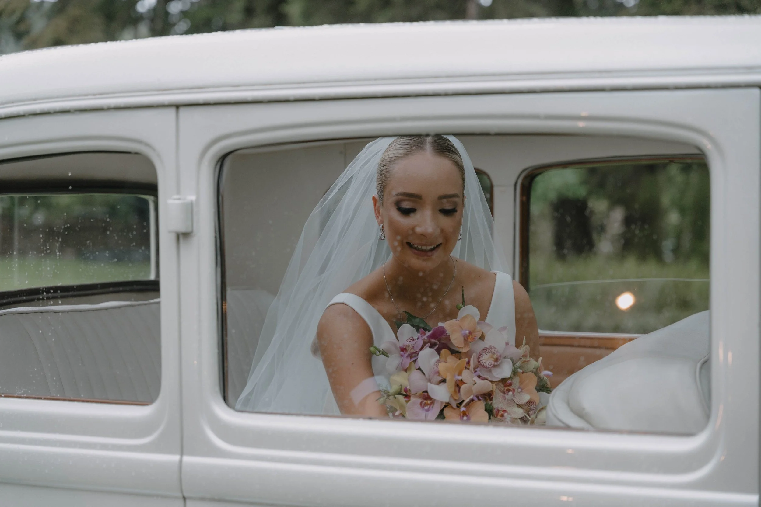 Mujer con vestido de novia y velo, sonriendo, sosteniendo un ramo de flores, dentro de un automóvil clásico blanco.