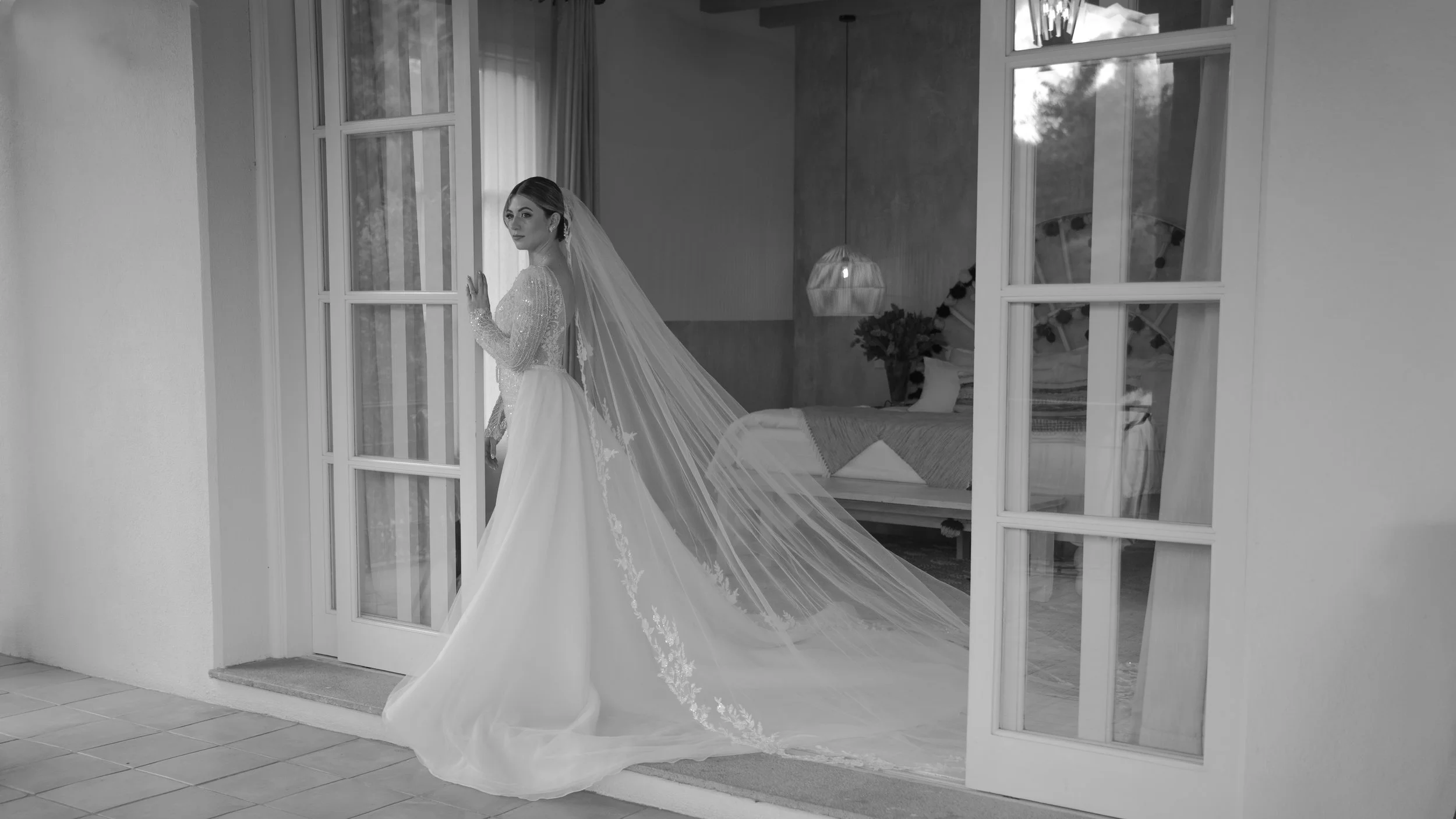 Mujer en vestido de novia con velo largo, mirando por una ventana en una habitación decorada con plantas y luz tenue.