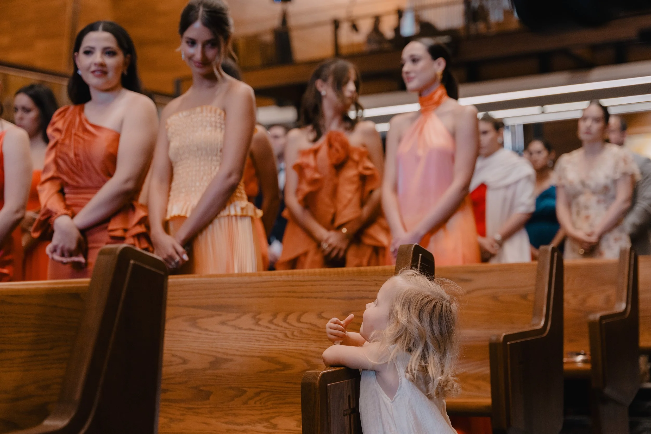 Niña con cabello rubio y largo mira hacia arriba, apoyada en el respaldo de una banca en una iglesia, mientras un grupo de mujeres vestidas con ropa elegante en tonos naranja y otros colores, están de pie al frente, participando en una ceremonia.