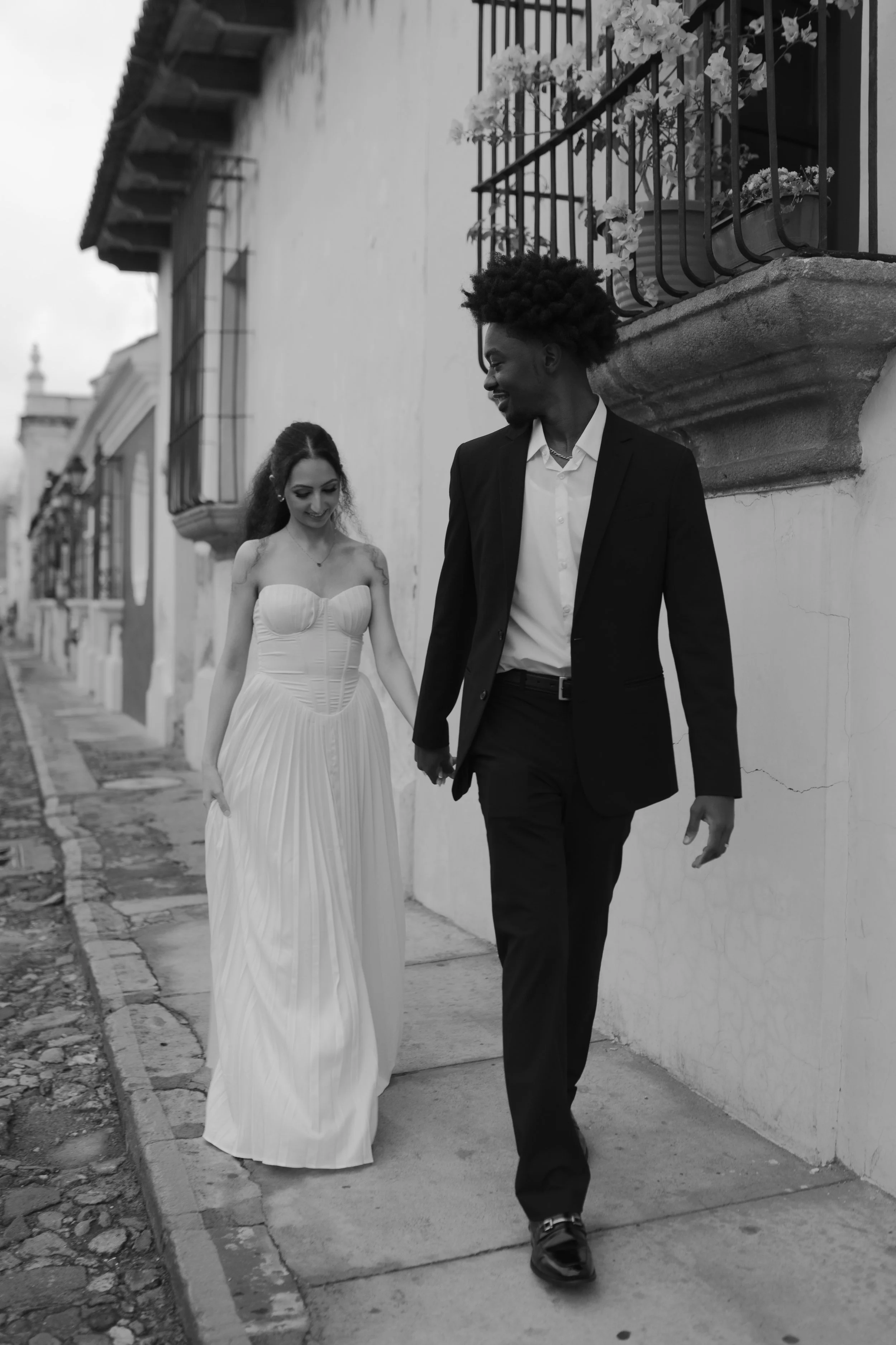 Pareja caminando juntos en la calle, la mujer lleva un vestido blanco y el hombre viste un traje negro.