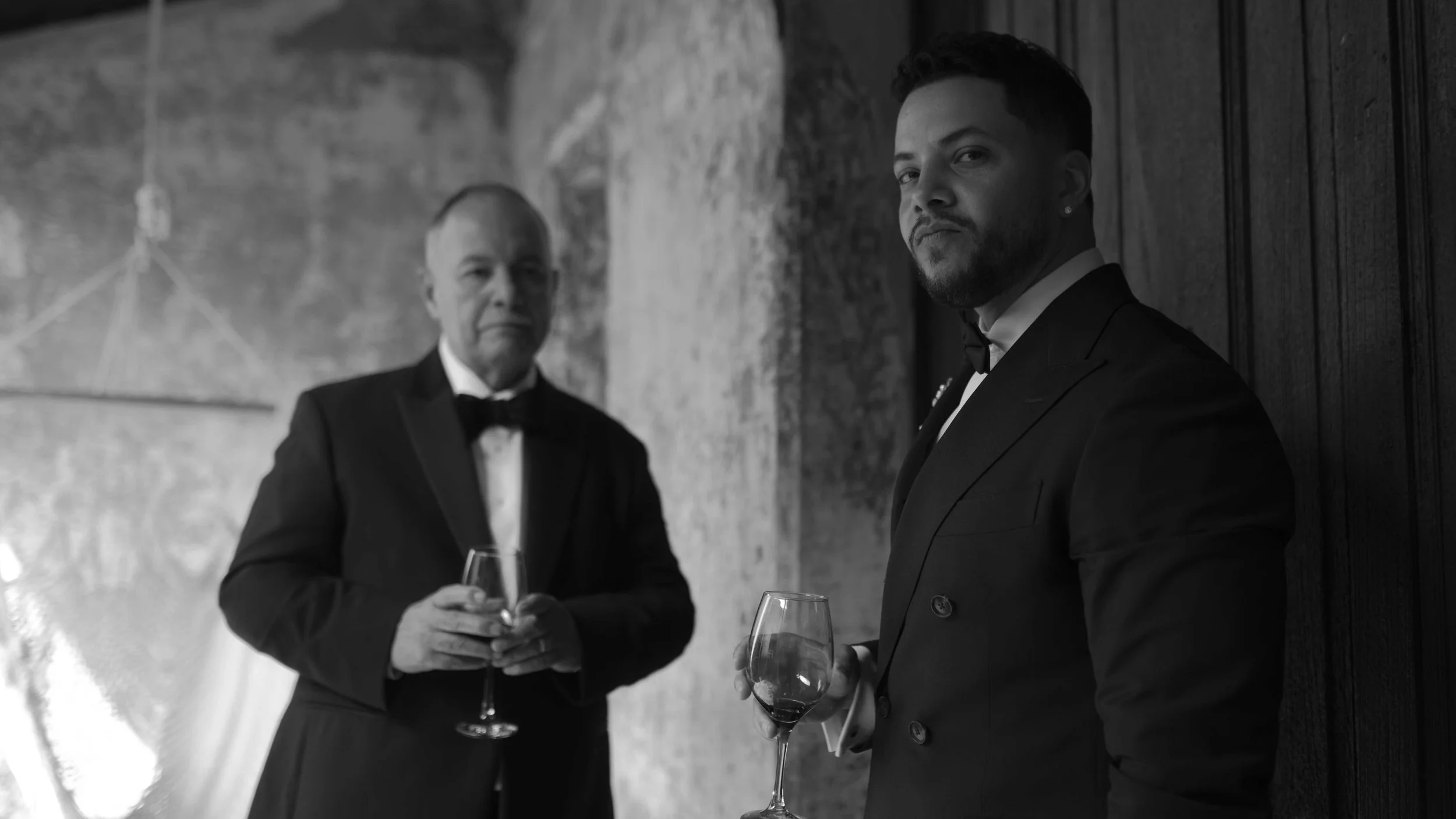 Dos hombres con trajes formales y corbatas, uno con barba, sosteniendo copas de vino en un ambiente elegante con paredes de piedra y madera.
