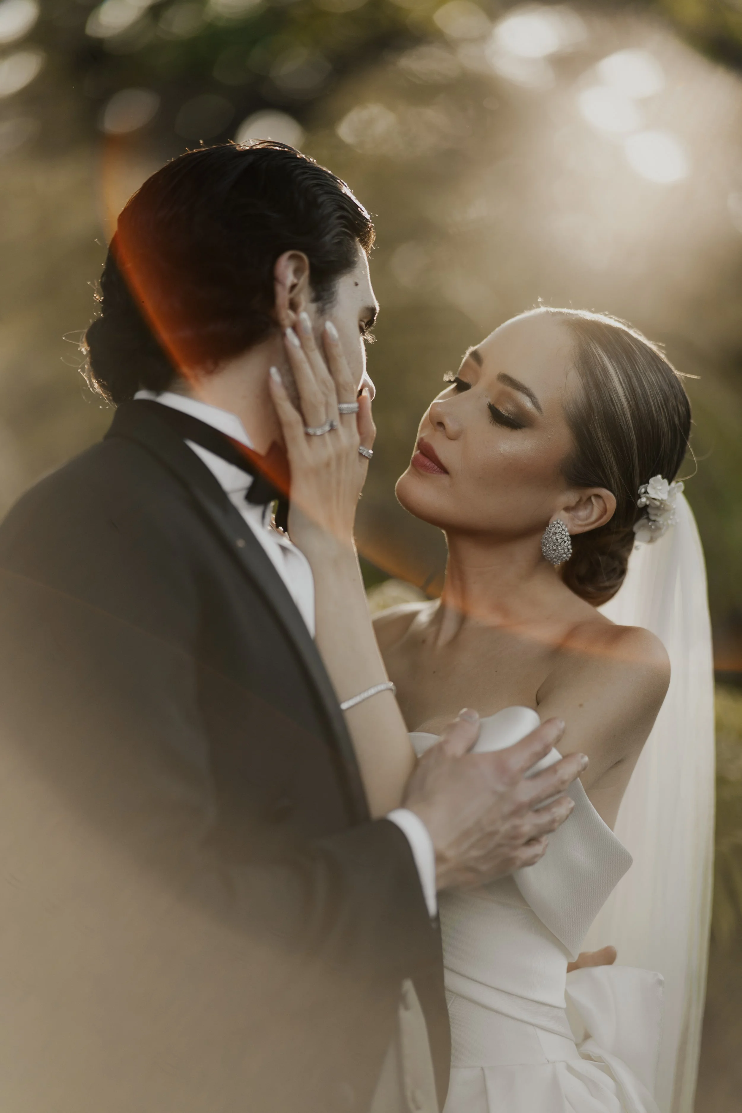 Una pareja de boda en un momento romántico, la mujer sostiene la cara del hombre con ambas manos en un entorno al atardecer.