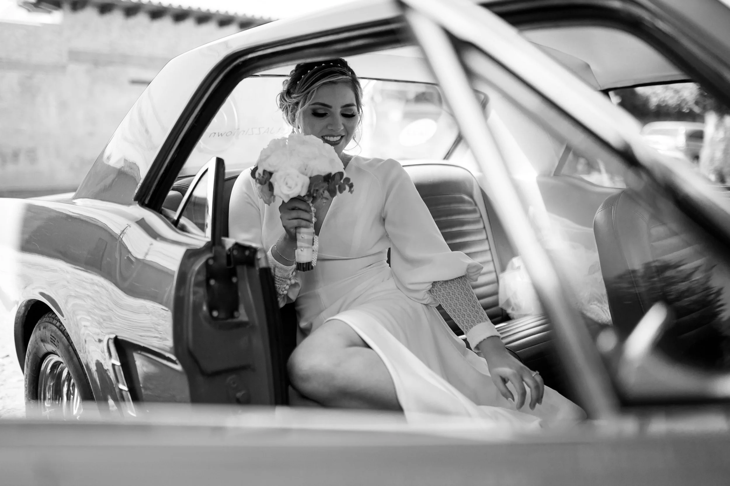 Mujer vestida de novia sentada en el asiento trasero de un coche clásico, sosteniendo un ramo de flores y sonriendo.