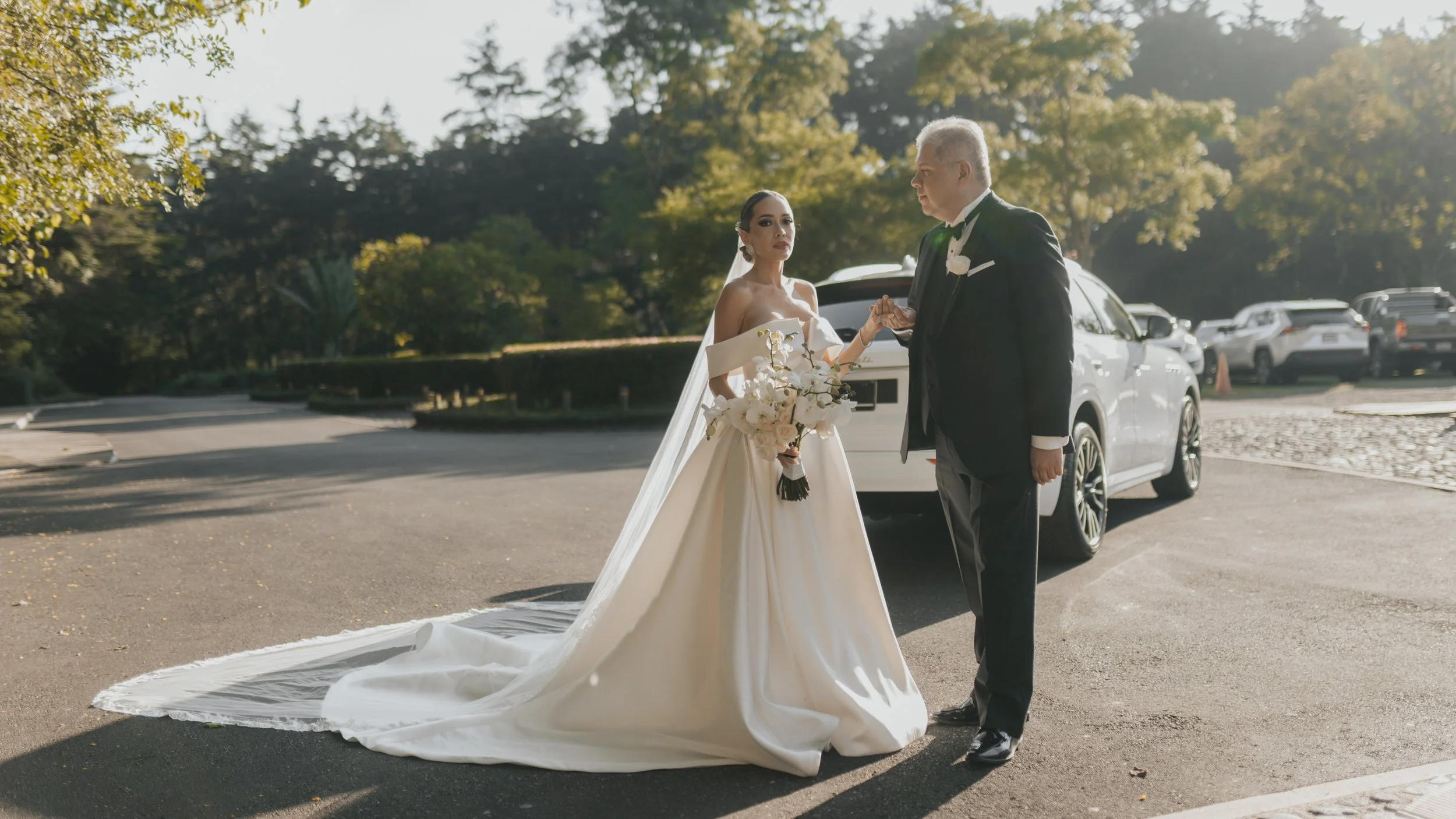 Pareja de novios en boda al aire libre, la novia con vestido de novia y ramo de flores, el novio en traje y corbata, sosteniendo la mano de la novia frente a un coche blanco, rodeados de árboles y autos.