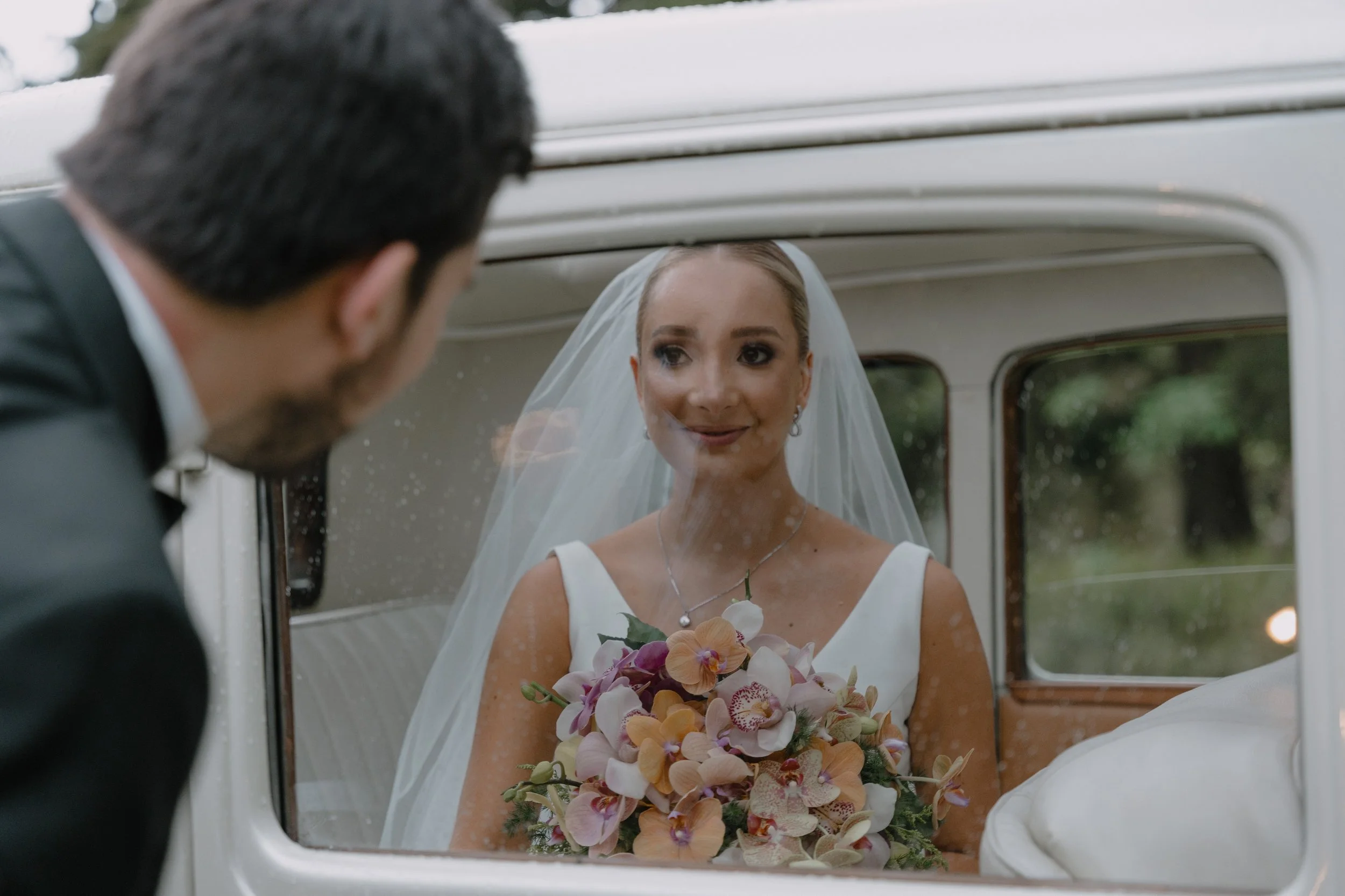Una novia con velo y vestido blanco observando a un hombre con traje, mientras mira por la ventana de un coche clásico y sostiene un ramo de flores