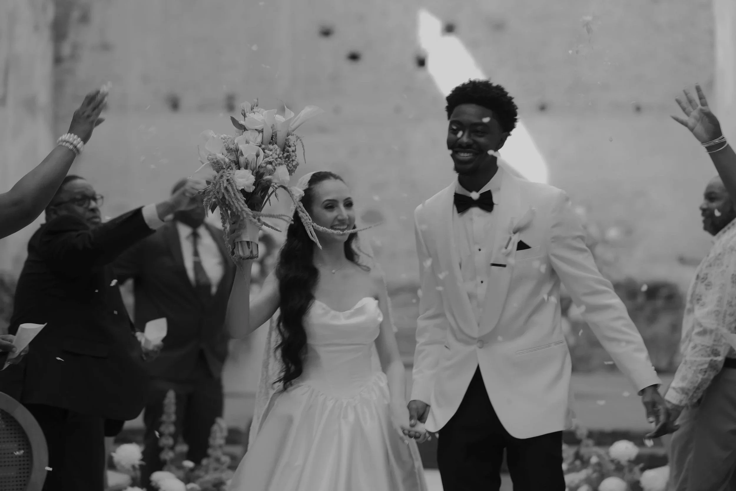 Una pareja de novios en una ceremonia de boda, ella sostiene un ramo de flores y se están dando la mano, rodeados de invitados en un lugar abierto.