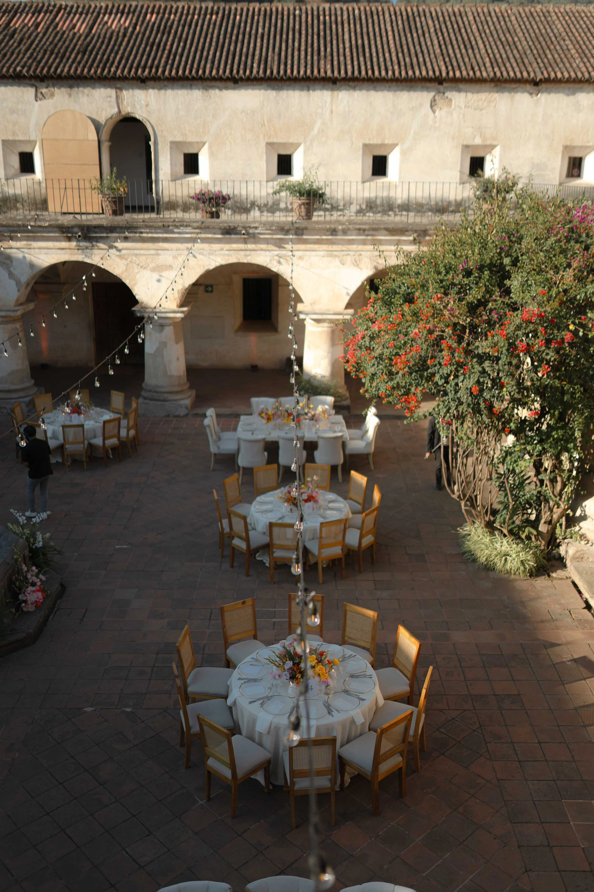 Decoración para un evento al aire libre con mesas redondas vestidas con manteles blancos y arreglos florales, en un patio con columnas, arbustos y un árbol con flores rojas, bajo cadenas de luces y fachada antigua en el fondo.