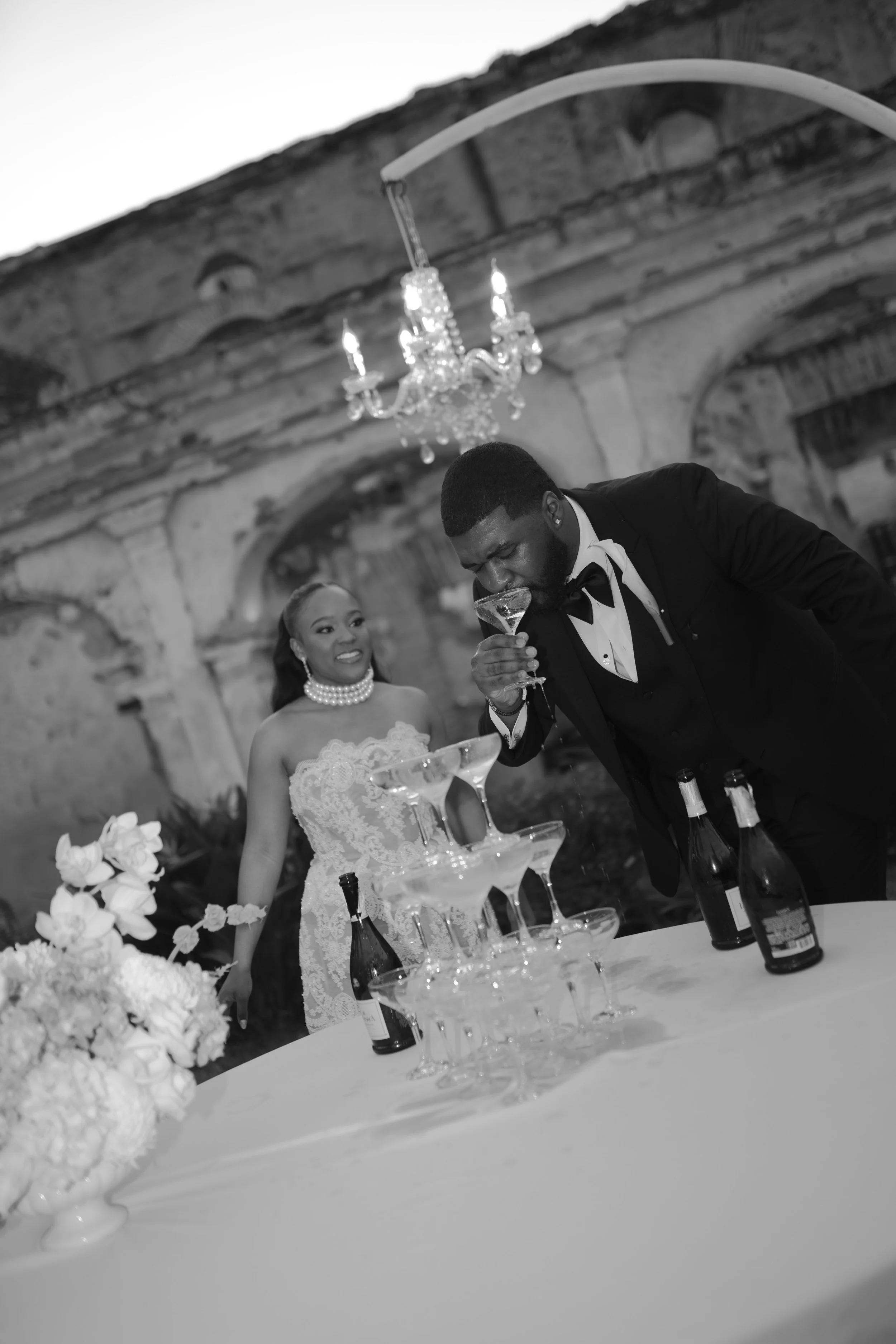 Pareja de novios en boda, el novio vierte una copa de champán, decoración con flores y una torre de copas en la mesa, en un lugar con techo de estructura antigua y un candelabro colgante.