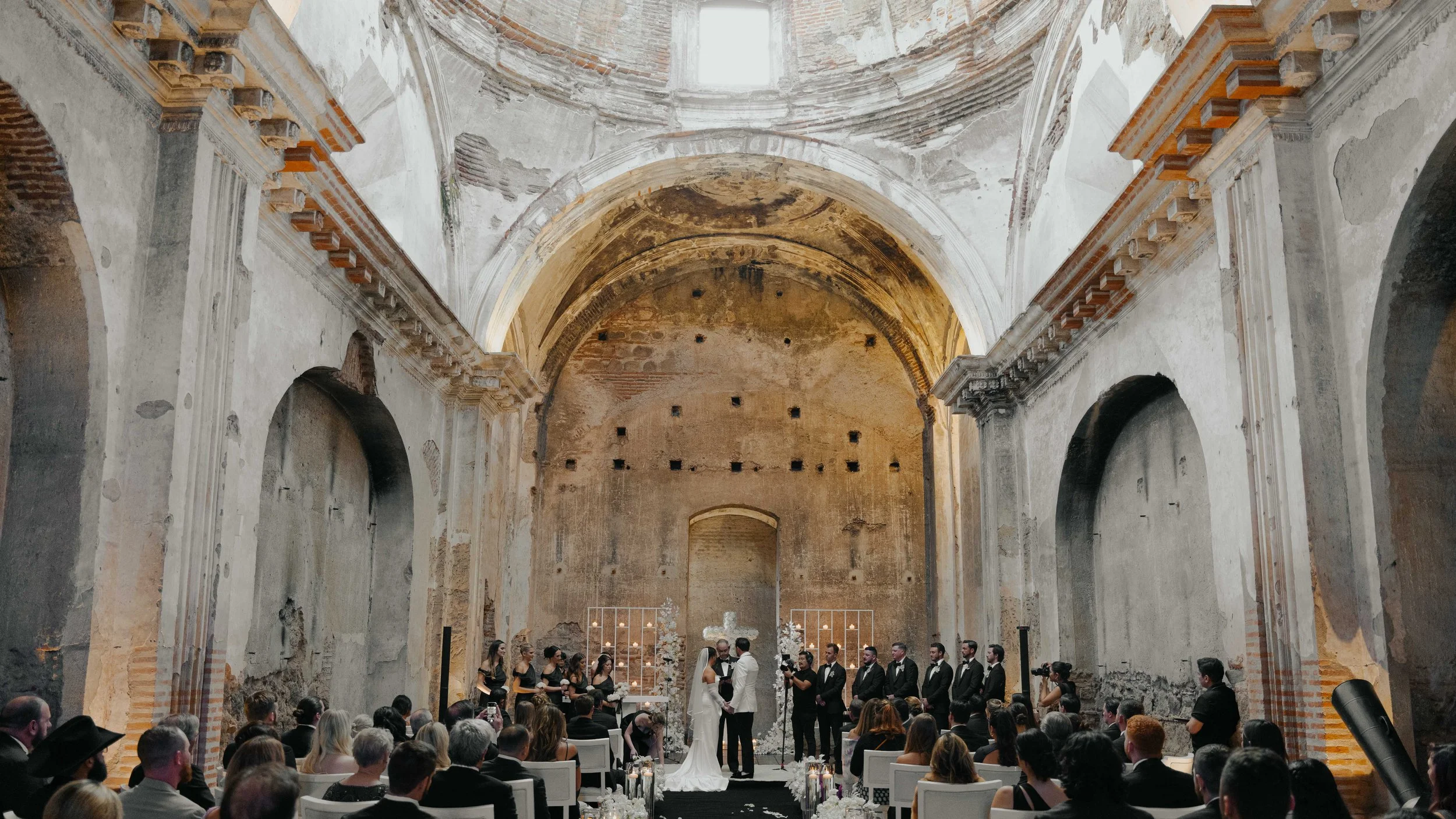 Boda en un antiguo templo con paredes desgastadas y techo en arcos, where una pareja intercambia votos frente a un altar floral, rodeada de invitados sentados y un equipo de grabación