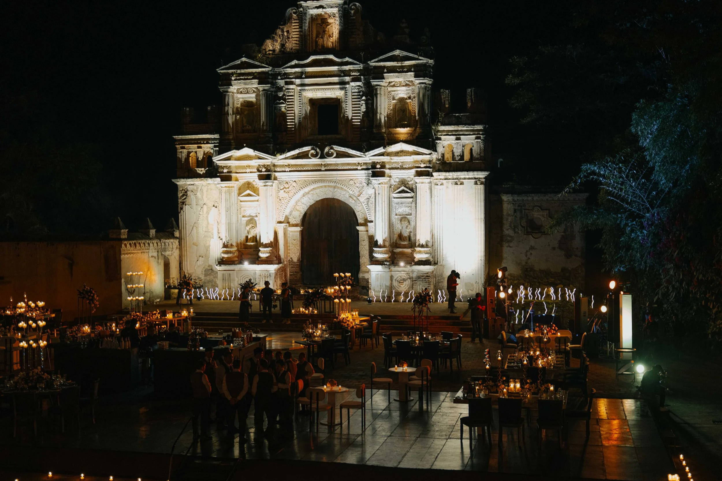 Celebración nocturna en la plaza con un edificio antiguo iluminado al fondo y mesas decoradas con velas en primer plano