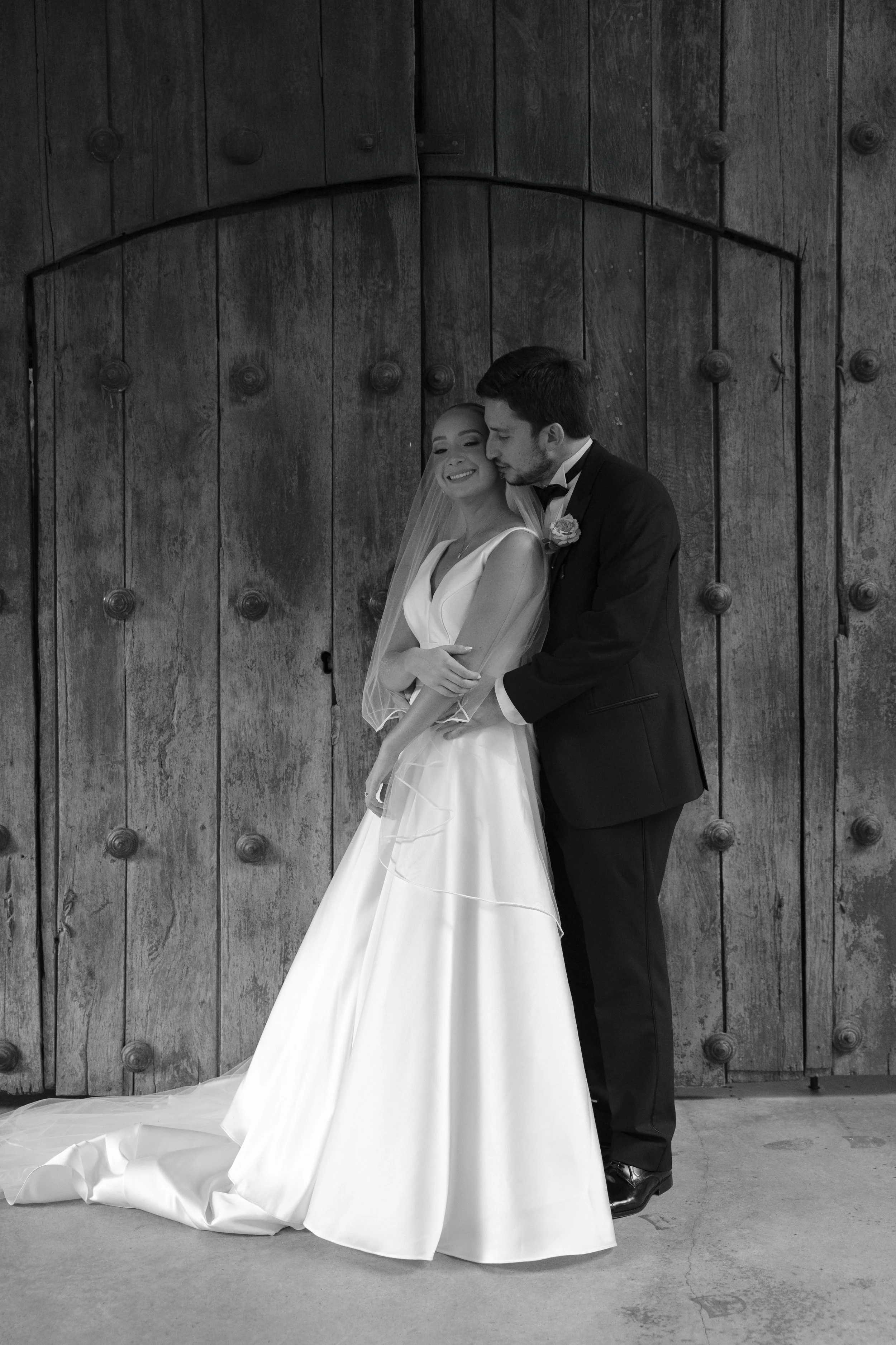 Pareja de recién casados en boda, la mujer con vestido de novia y el hombre con traje negro, en un fondo de puerta de madera.