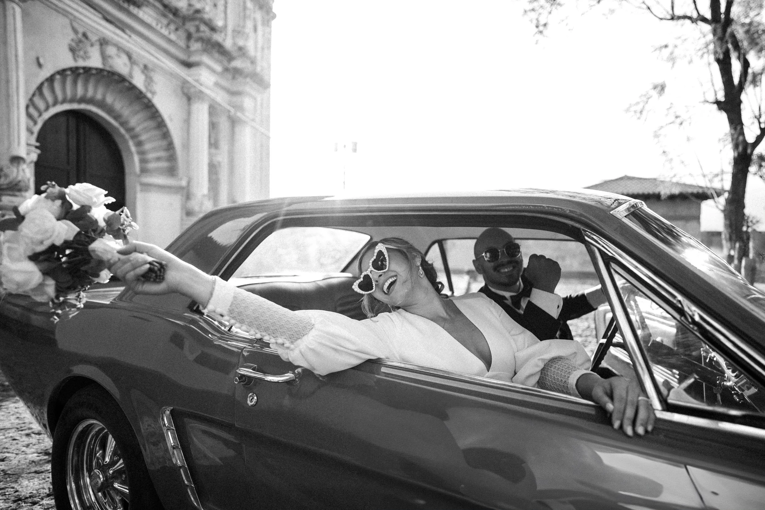 Pareja de novios en un coche clásico, la novia sosteniene un ramo de flores y lleva gafas grandes con forma de corazones, el novio lleva gafas de sol y ambos sonríen felices, en un fondo de arquitectura antigua y árboles.