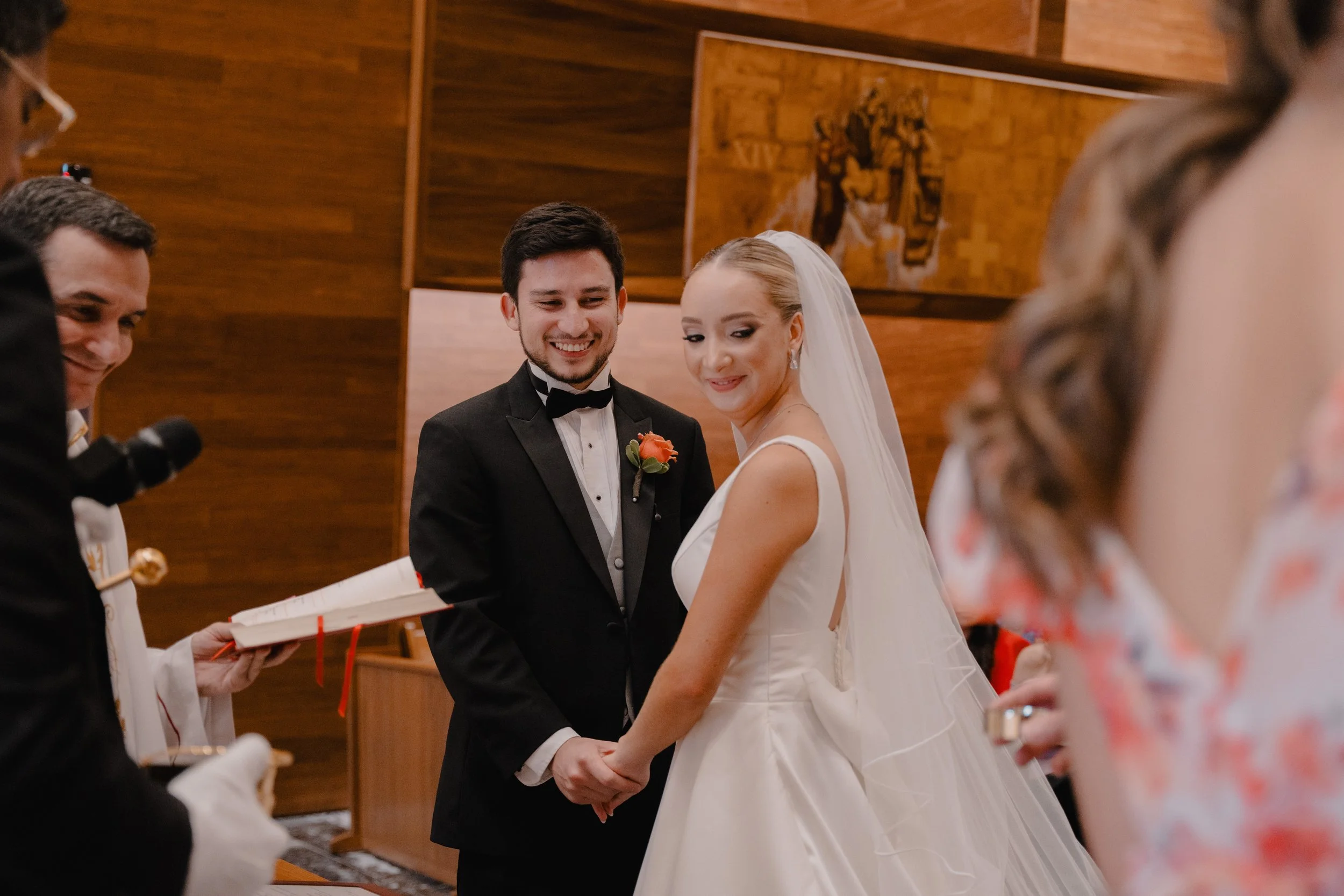 Pareja de novios en una ceremonia de boda, la novia lleva vestido blanco y velo, el novio en saco y pajarita, sosteniéndose de las manos y sonriendo. Hay personas a su alrededor en un ambiente elegante de madera.