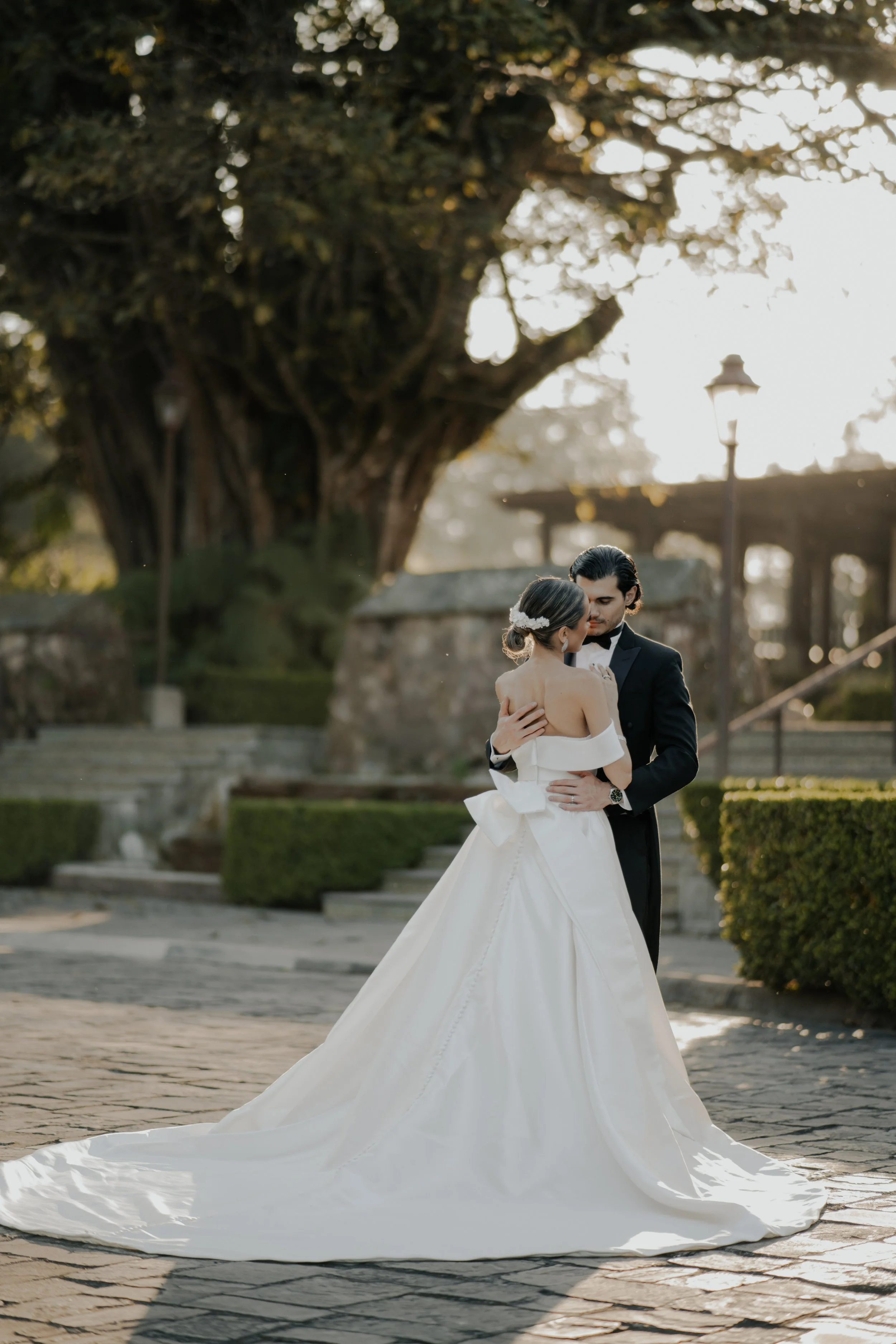 Pareja de novios bailando en un ambiente al aire libre, con fondo de naturaleza y un árbol grande, durante el atardecer.