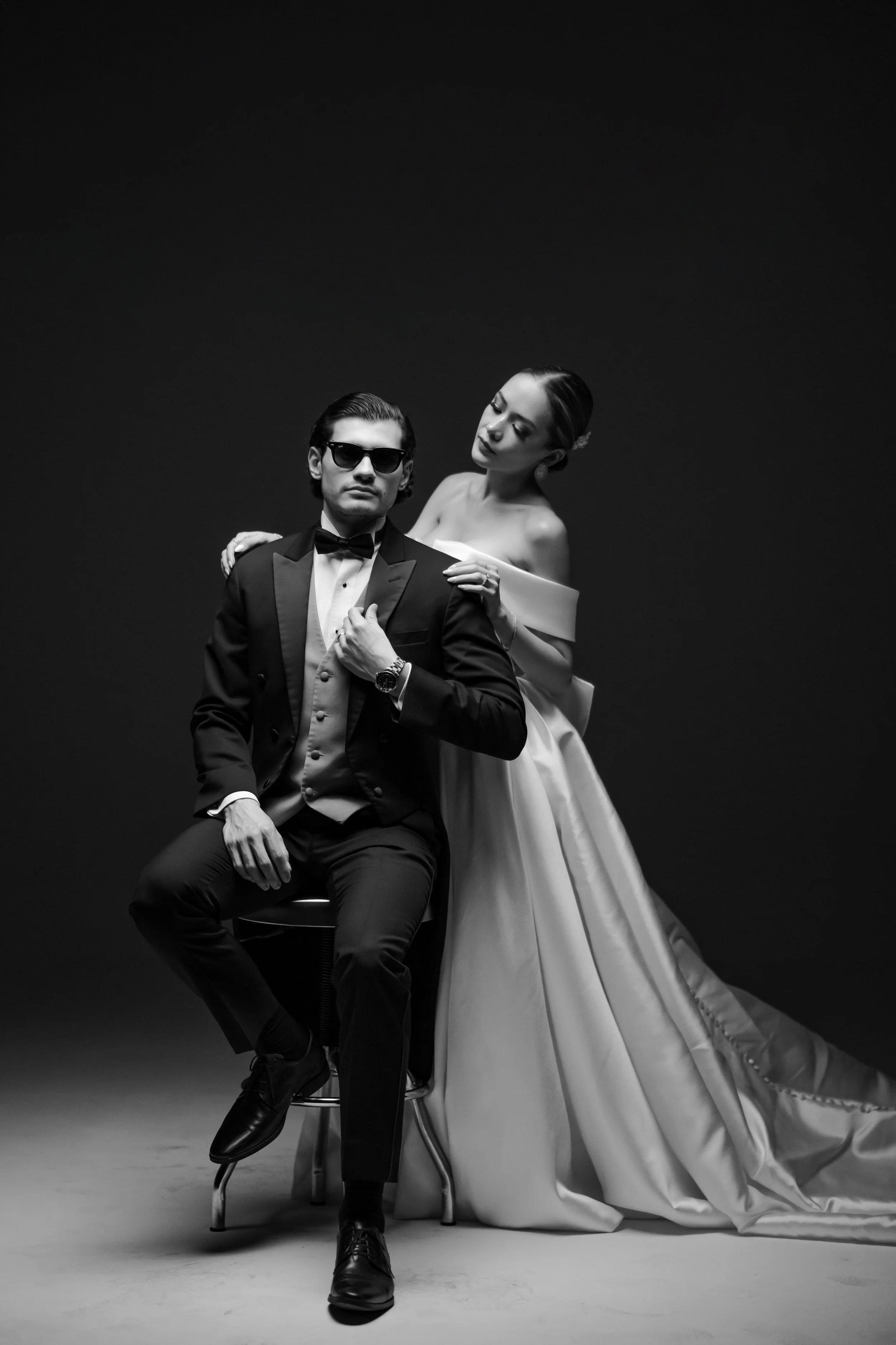 Foto en blanco y negro de una pareja elegante, con el hombre con disfraz de tuxedo y gafas de sol, y la mujer con vestido de novia, en un estudio con fondo oscuro.