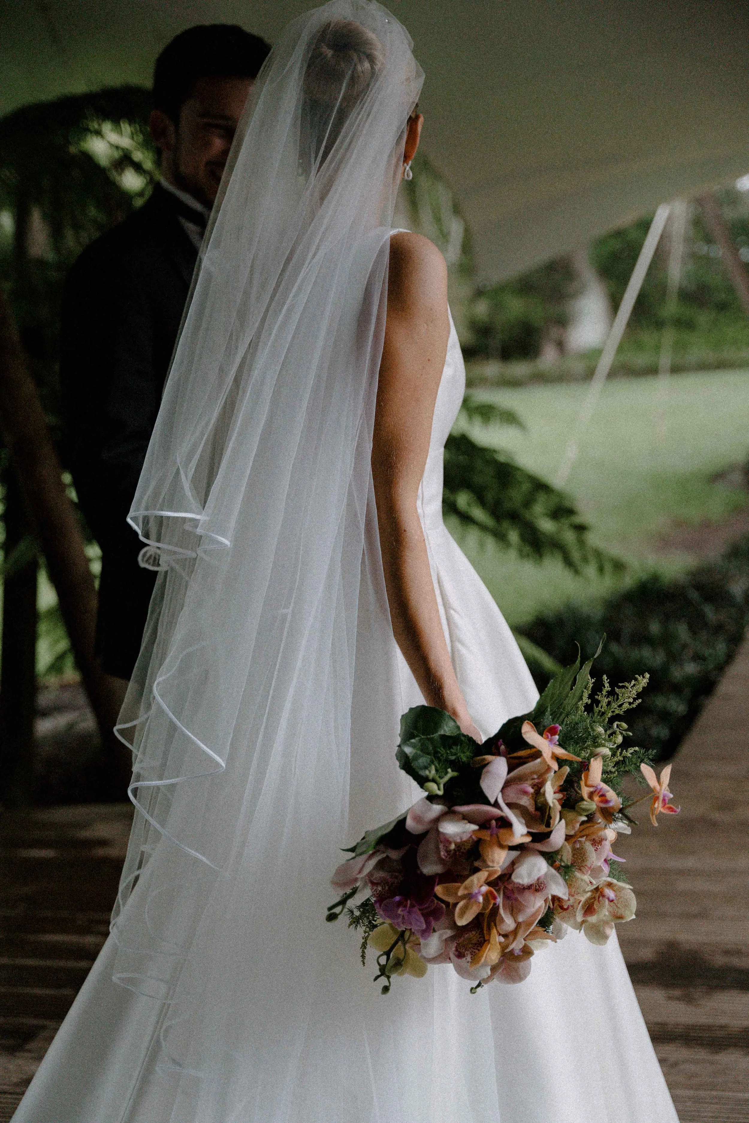 Una novia vestida con un vestido de boda blanca y un velo largo, sosteniendo un ramo de flores en un entorno natural, con un fondo de árboles y césped.