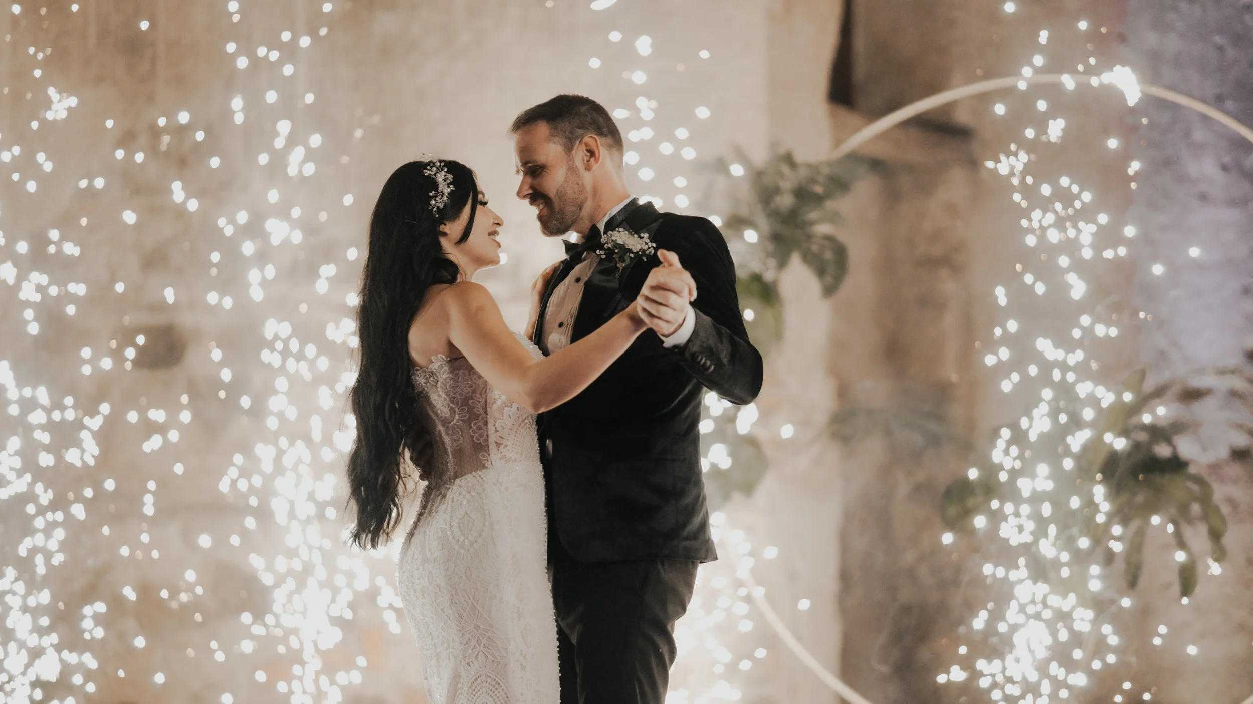Pareja bailando en su boda, rodeada de luces decorativas y fondo de piedra.