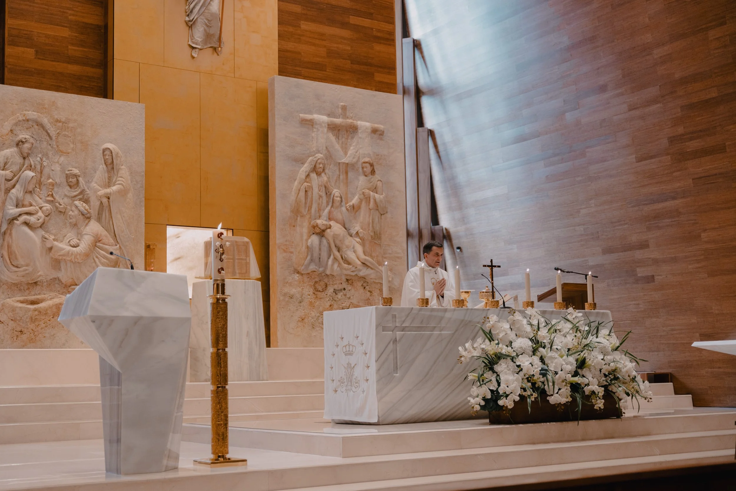 Un sacerdote en una iglesia realizando una ceremonia. La mesa del altar está decorada con flores blancas y tiene cinco cirios encendidos. Hay esculturas religiosas en el fondo y el ambiente es de oración y reverencia.