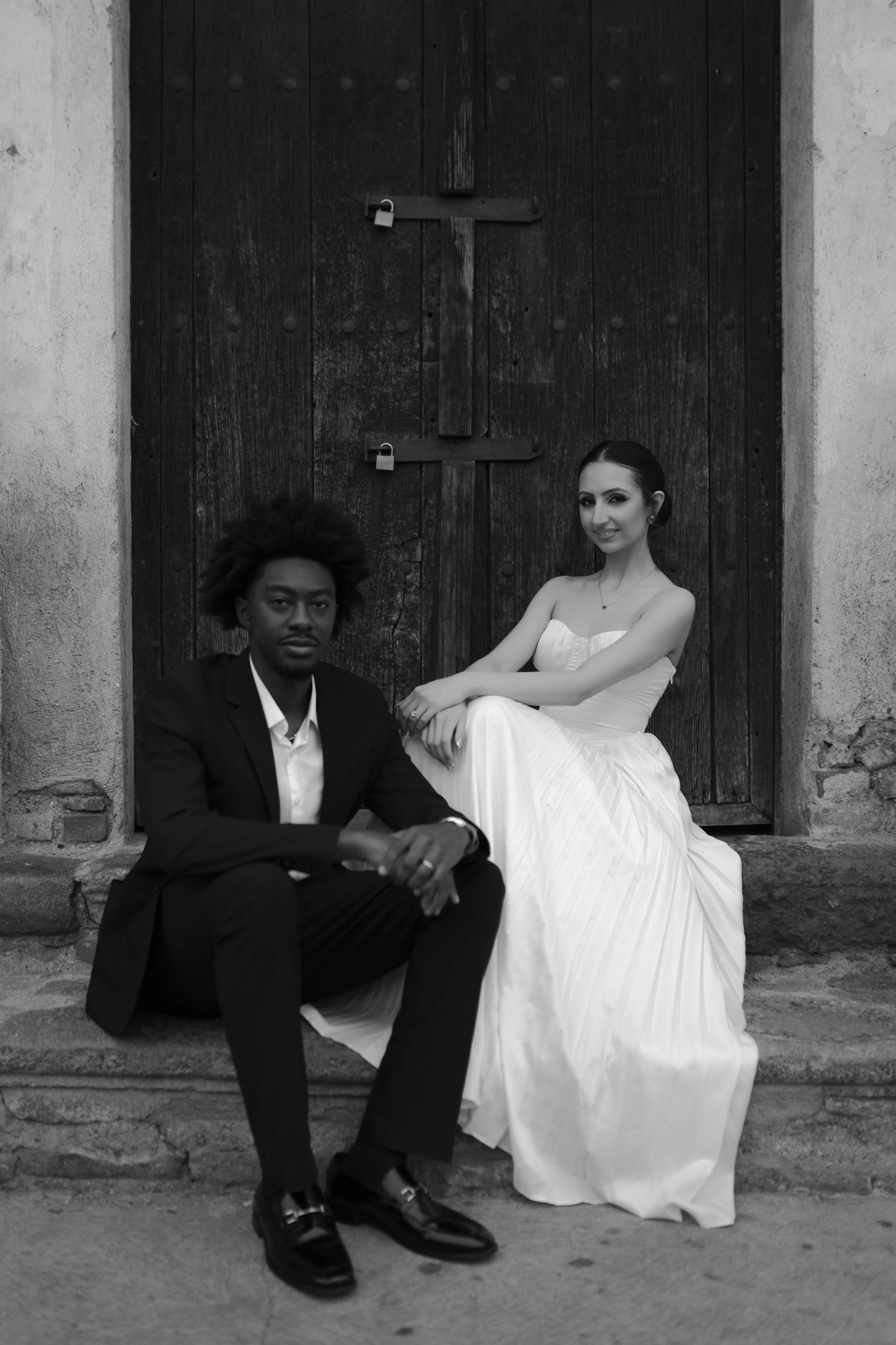 Una pareja vestida con ropa elegante, uno con traje negro y la mujer con vestido blanco, sentados sobre un escalón frente a una puerta de madera, en una imagen en blanco y negro.