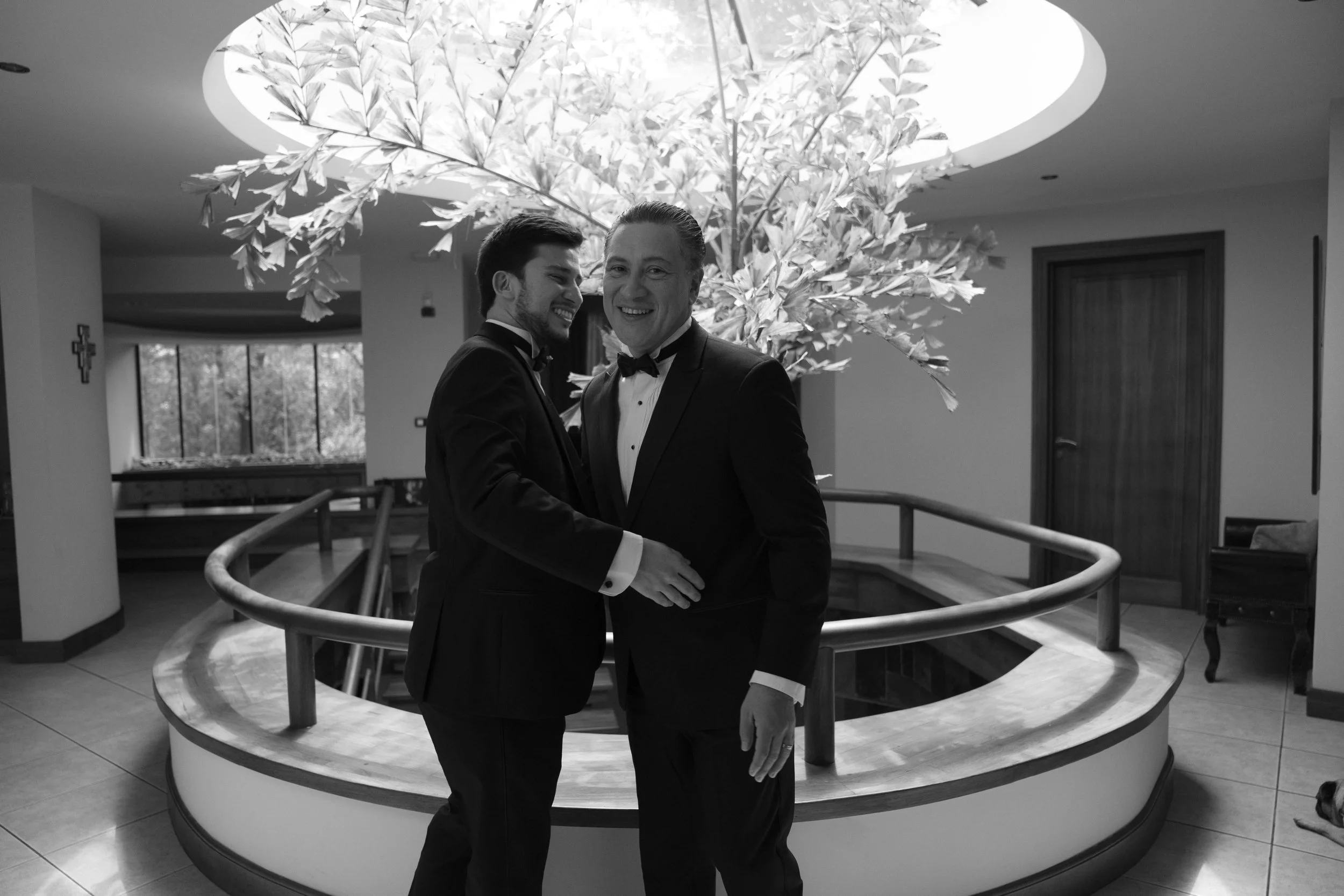 Dos hombres vestidos con trajes de etiqueta, uno más joven y otro de mediana edad, abrazándose y sonriendo en un interior moderno con una iluminación artística en forma de árbol en el techo.