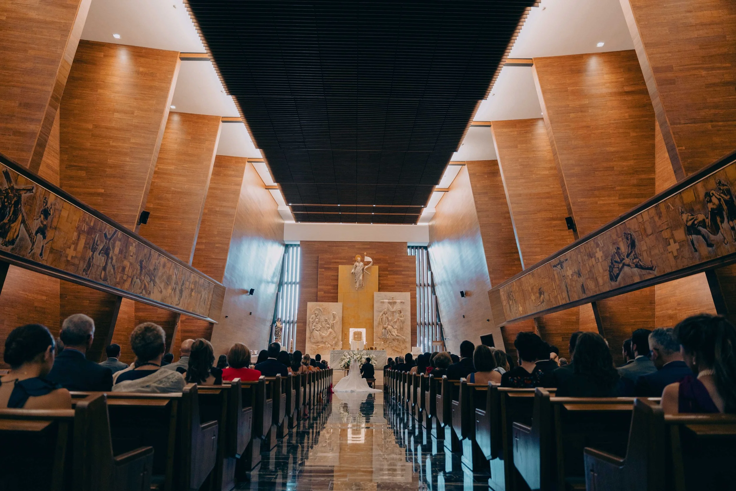Interior de una iglesia o capilla con asistentes en una ceremonia, vista desde el suelo hacia el altar con un crucifijo y esculturas religiosas, paredes de madera y ventanas altas con cortinas.
