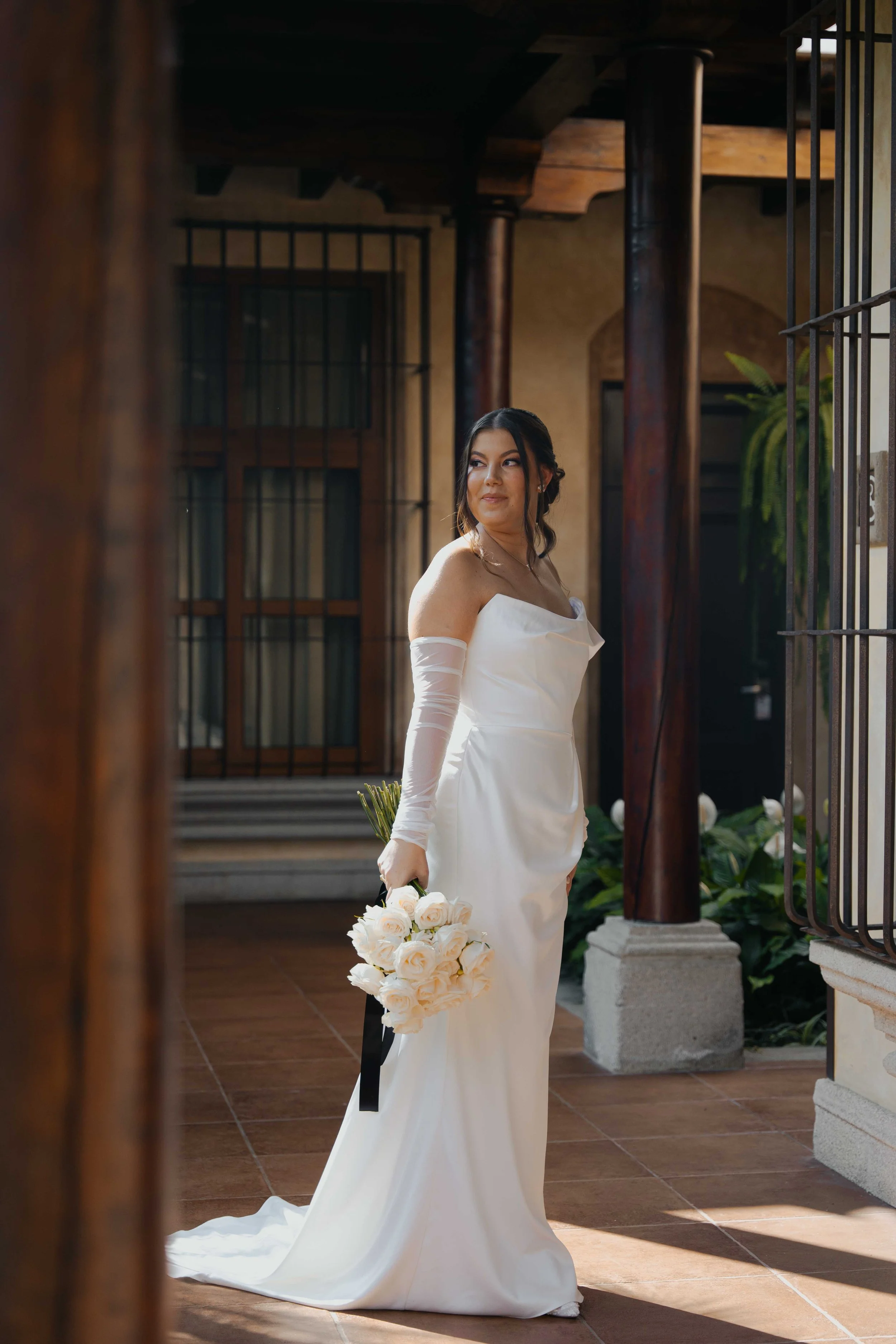 Mujer en vestido de novia sosteniendo un ramo de rosas blancas en un espacio con columnas de madera y barrotes de hierro.