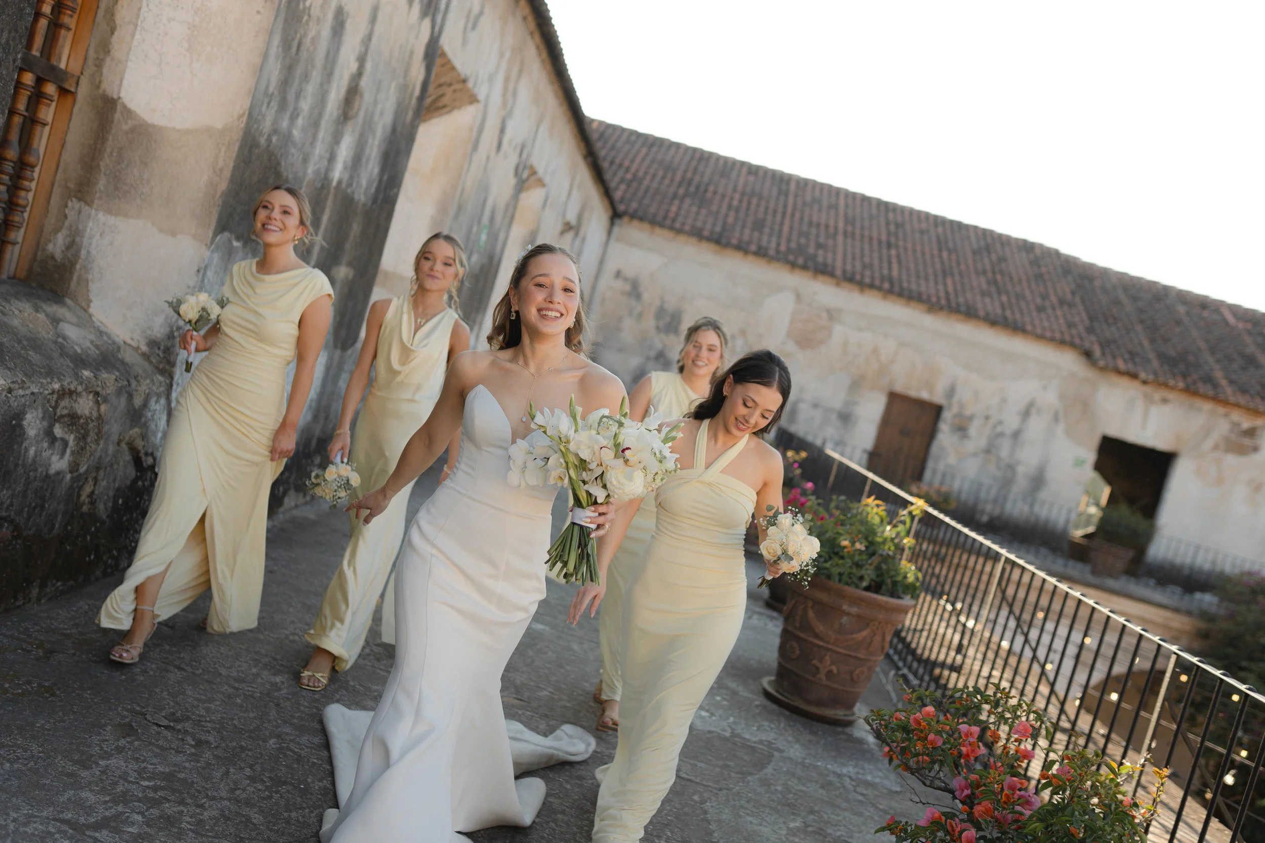 Grupo de seis mujeres, una de ellas en vestido de novia, caminando felices en un patio con paredes de piedra y techo de tejas durante una boda.