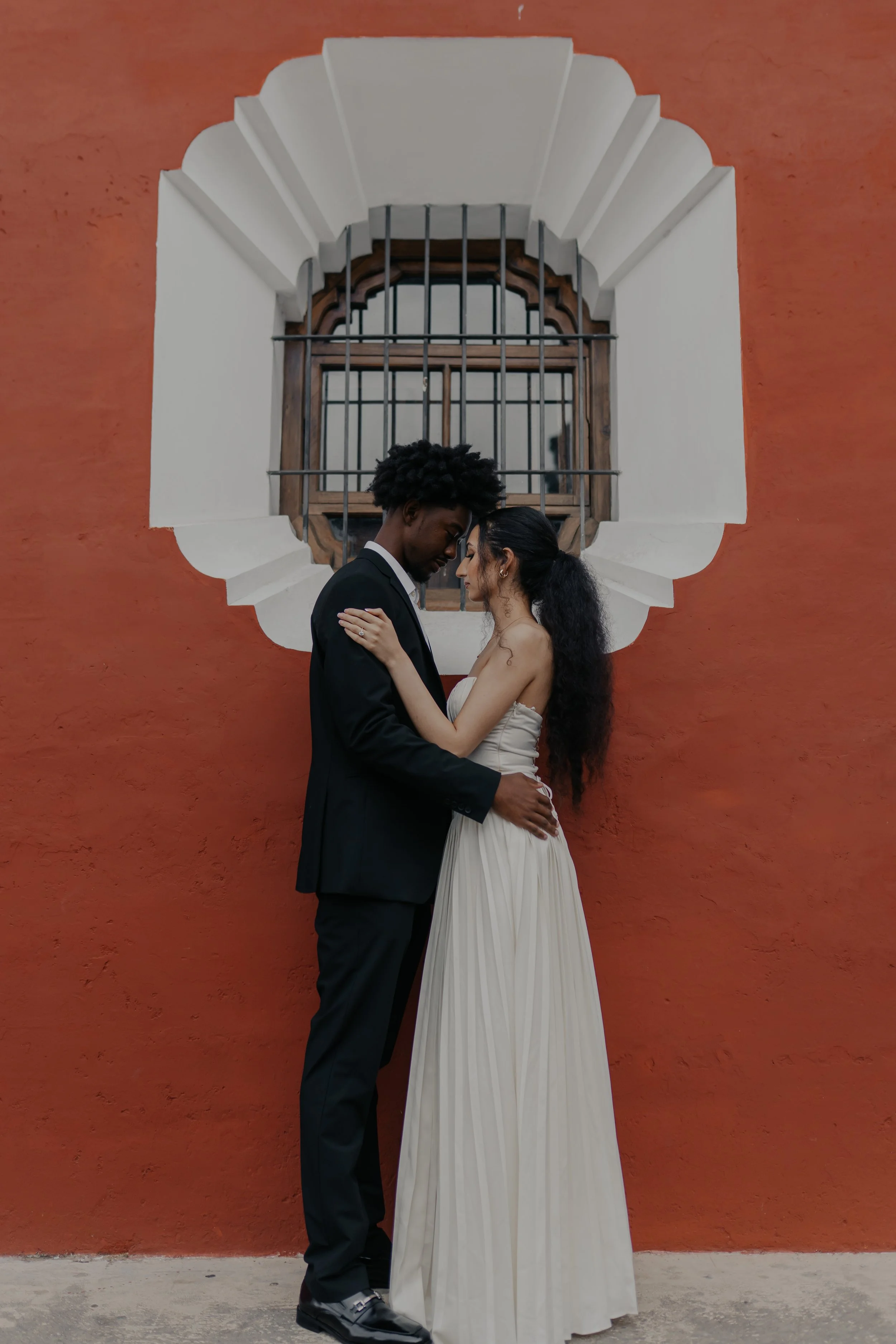 Una pareja vestida de novia y novio se abraza frente a una pared de color terracota con una ventana de madera y rejas de metal.