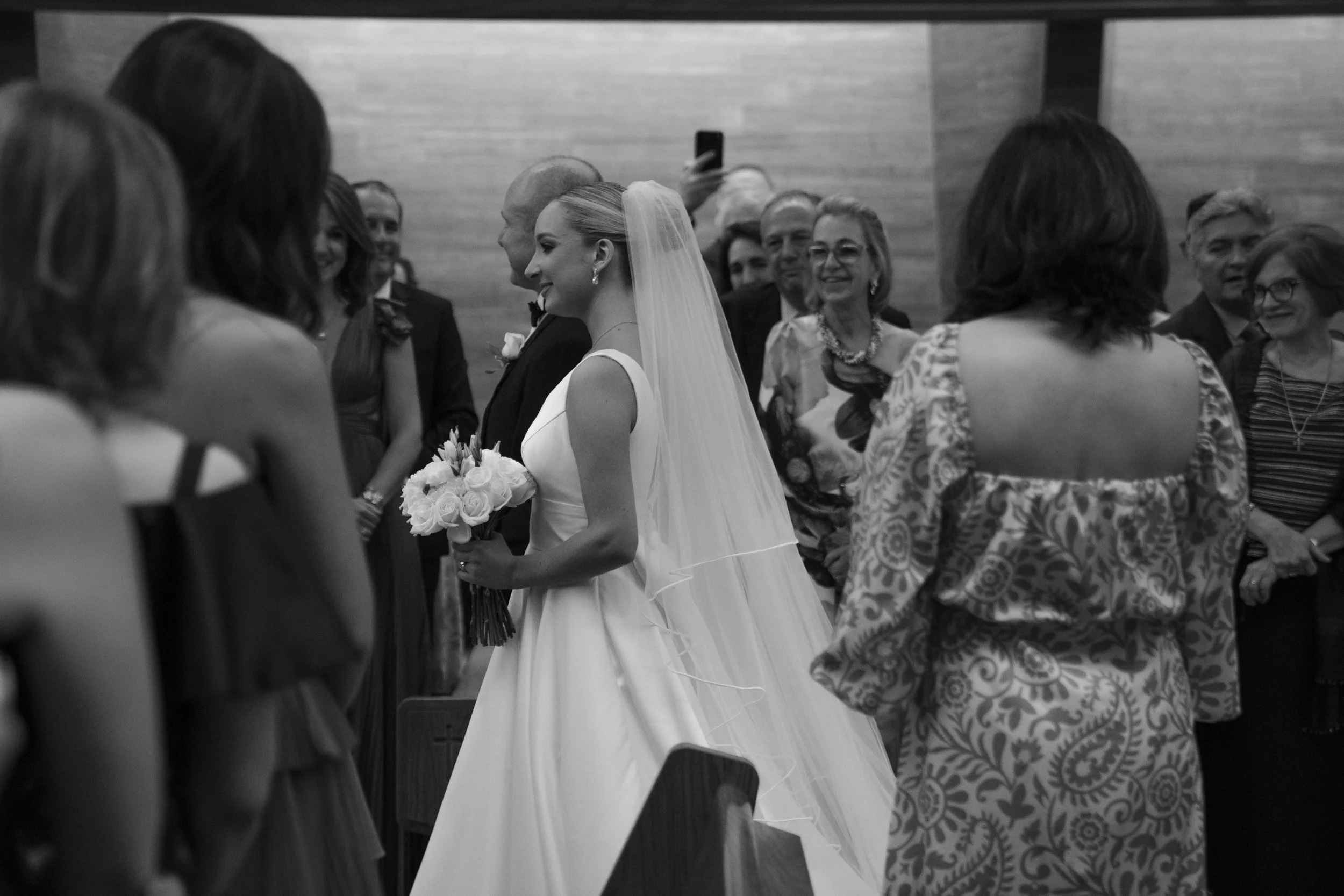 Mujer con vestido de novia y velo, sosteniendo un ramo de flores blancas, en una ceremonia de boda con varios invitados observando y sonriendo.