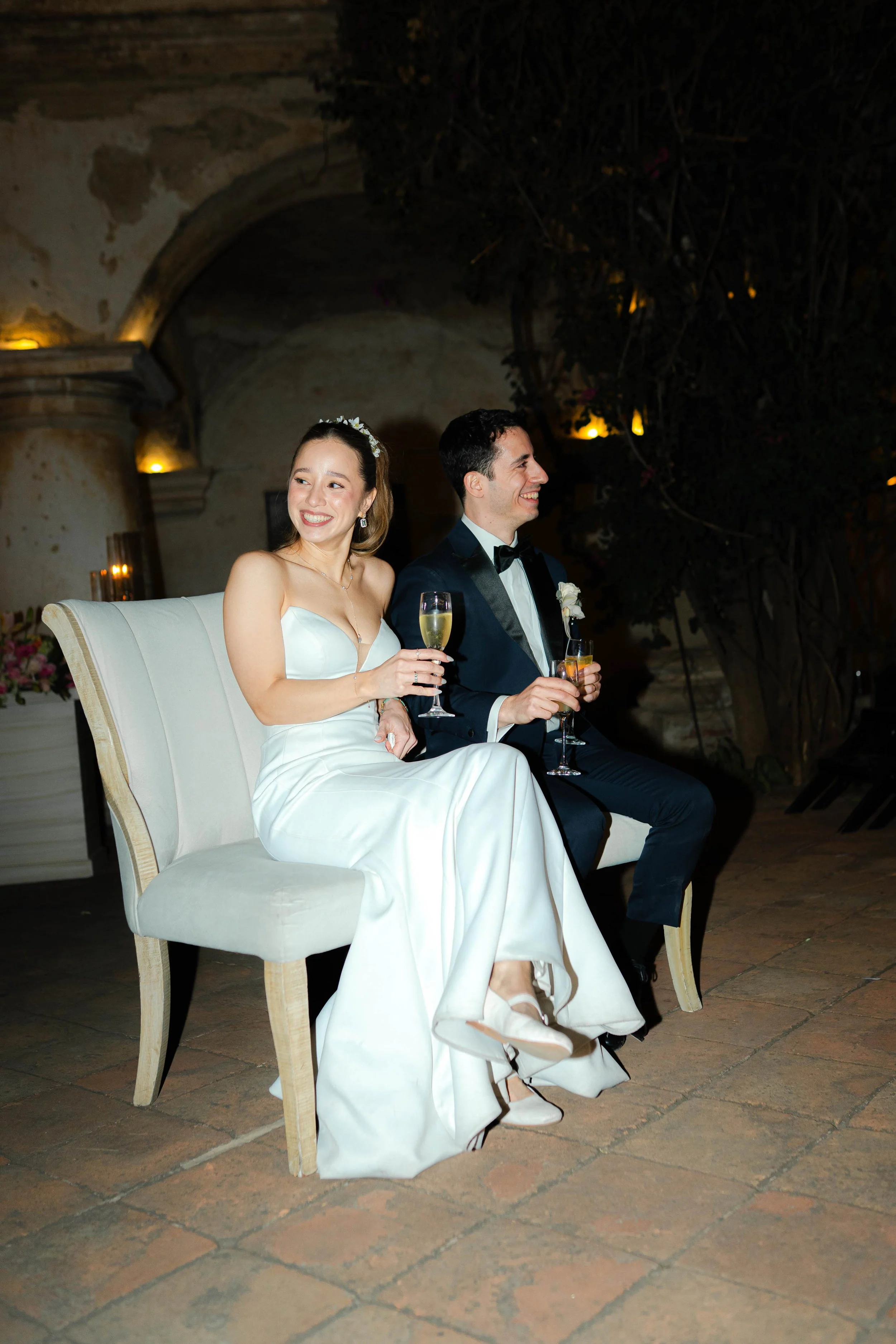 Pareja de recién casados sentados en un sofá, sonriendo y brindando con copas de champagne en una celebración.