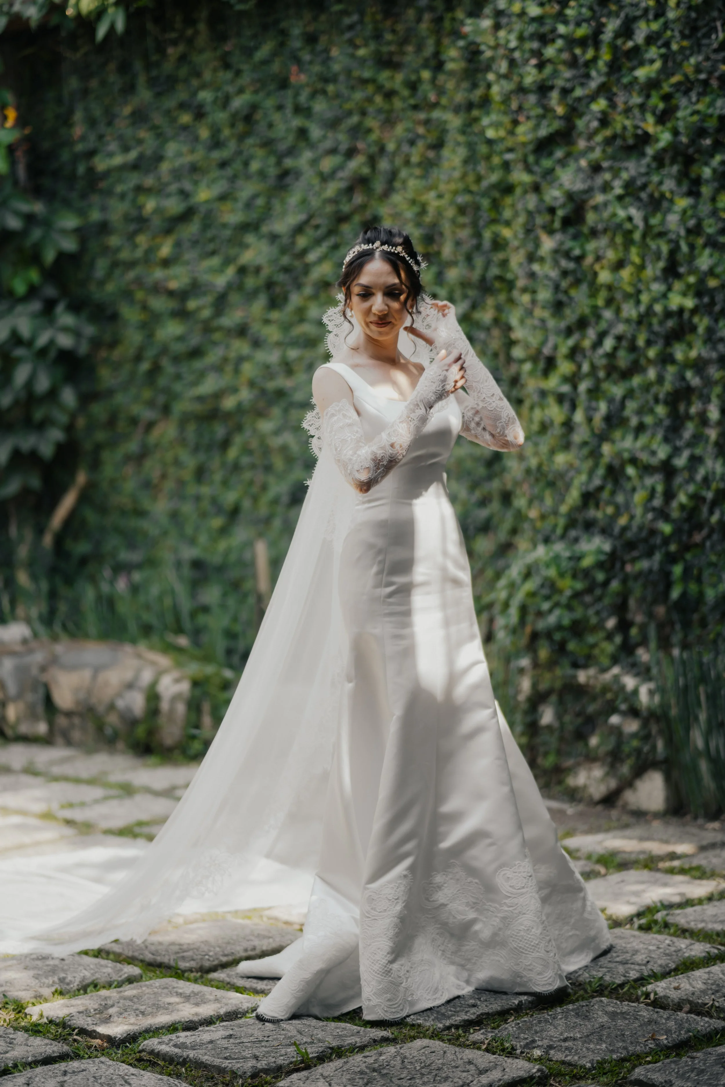 Una novia con vestido blanco largo y guantes de encaje, posando en un jardín con paredes verdes y piedras en el suelo.